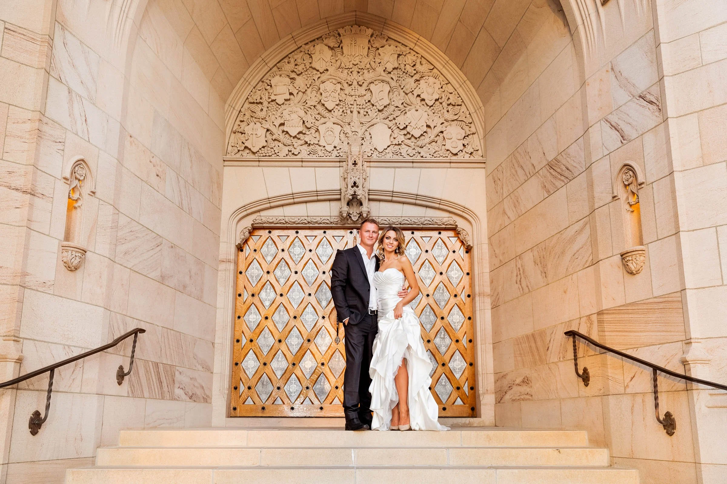 A bride and groom standing on the steps of a church or cathedral, with intricate carvings and a wooden lattice behind them.