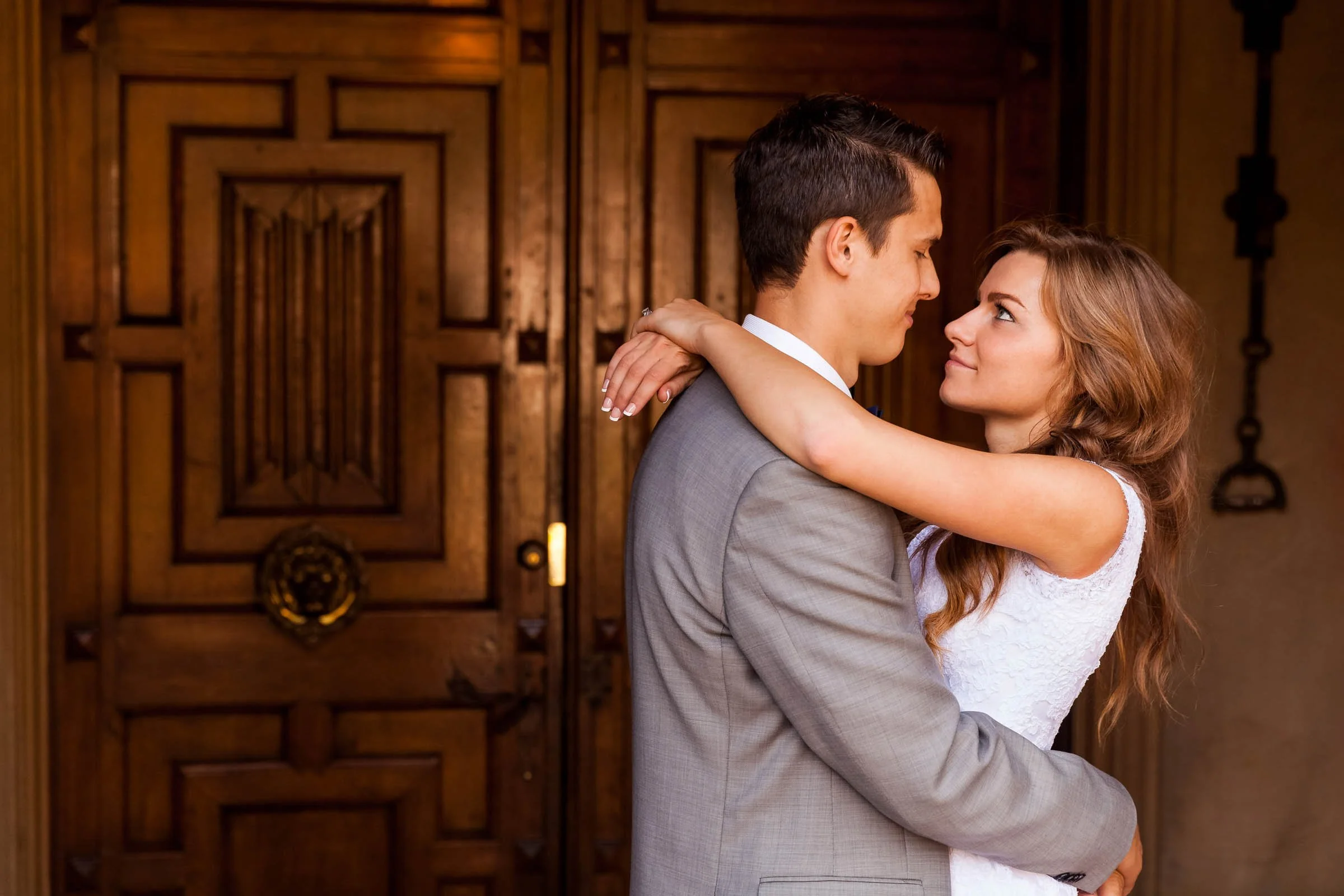 A bride and groom sharing a romantic moment inside a warmly lit room with intricate wooden doors in the background.