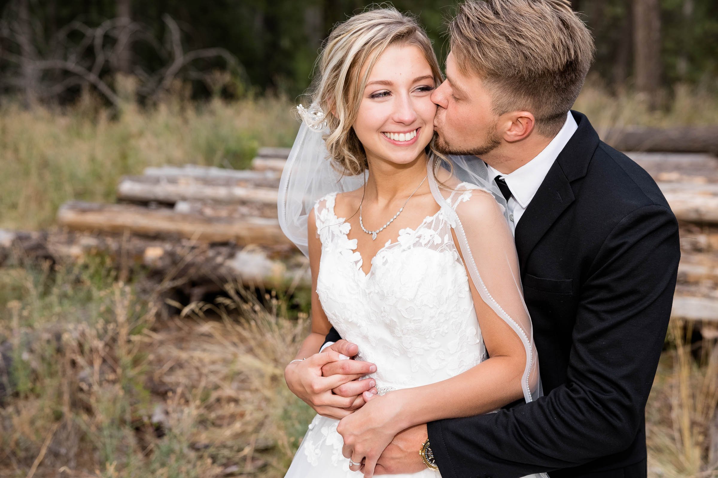 A smiling bride in a white wedding dress and a groom in a black suit sharing a kiss outdoors, with logs and trees in the background.