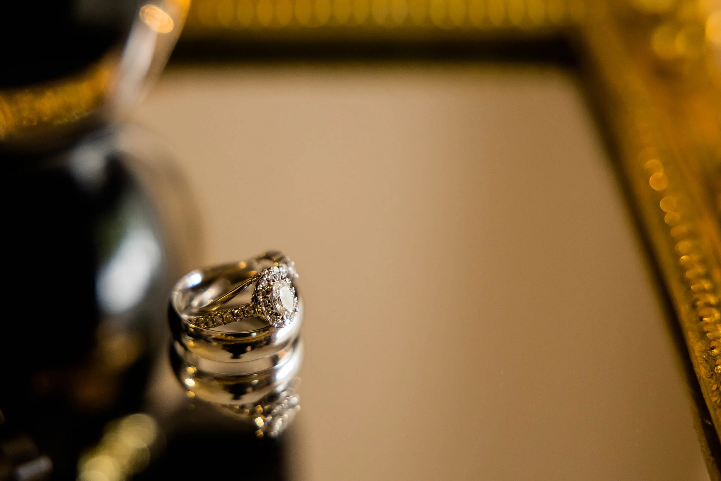 A close-up of a diamond engagement ring and wedding band on a reflective surface with a blurred mirror and gold frame in the background.