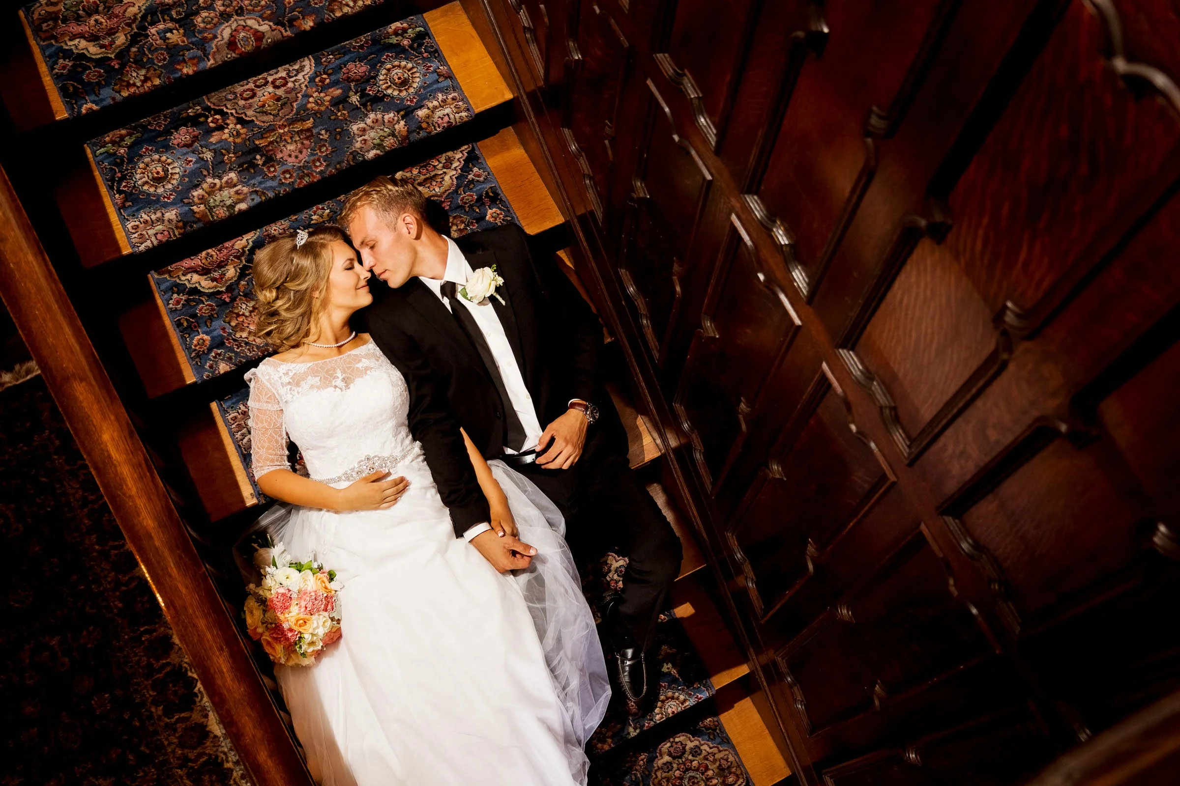 A bride and groom lying on a staircase, facing each other and holding hands, in wedding attire. The bride is in a white lace dress holding a bouquet, and the groom is in a black tuxedo with a white boutonniere.
