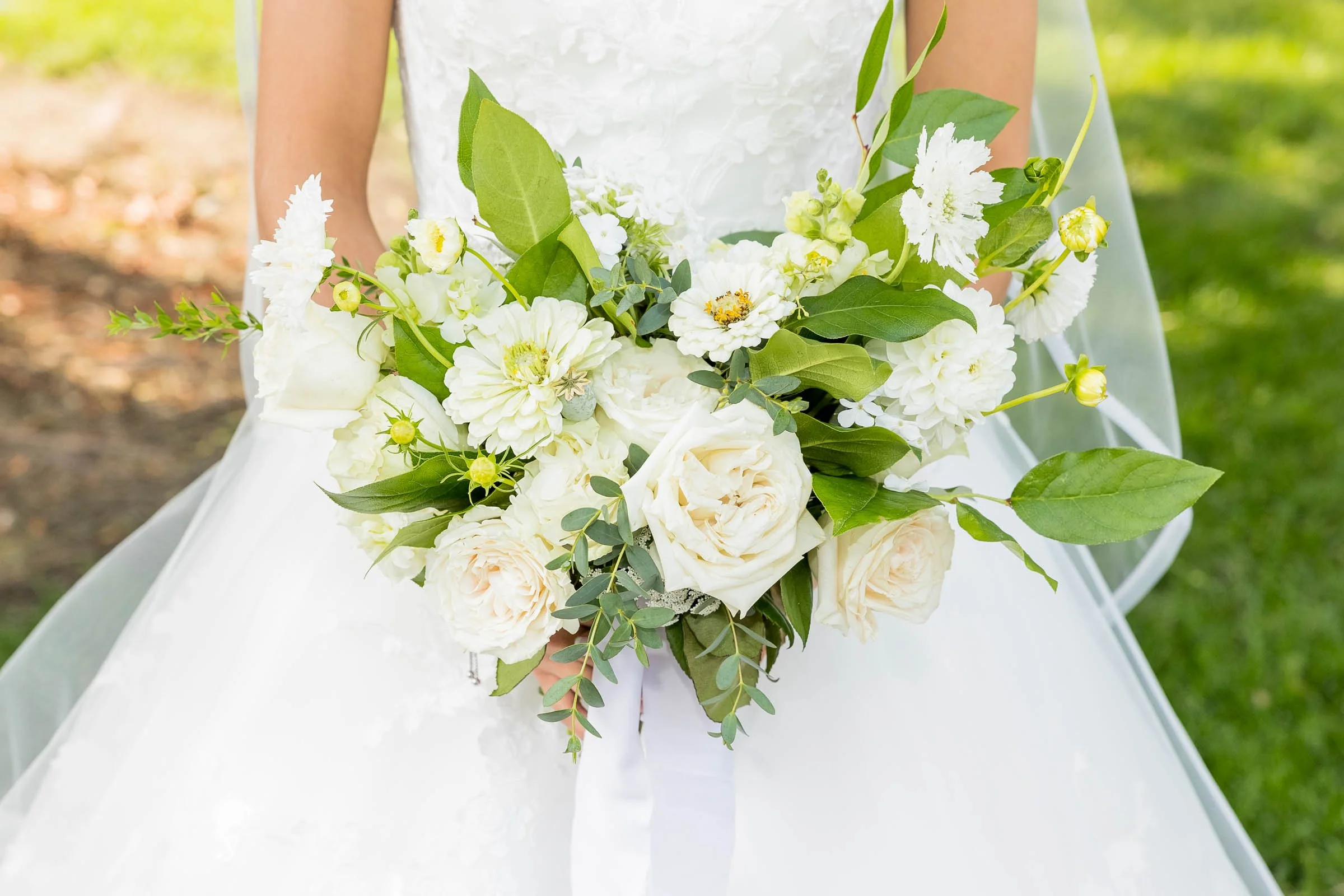 A bride holding a bouquet of white flowers and green leaves outdoors on grass.