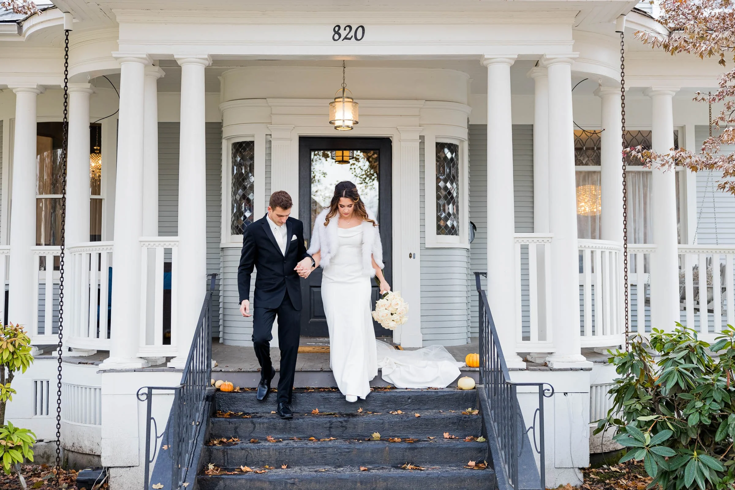 A bride and groom descending the steps of a house, holding hands, with pumpkins on the stairs, on a fall day.