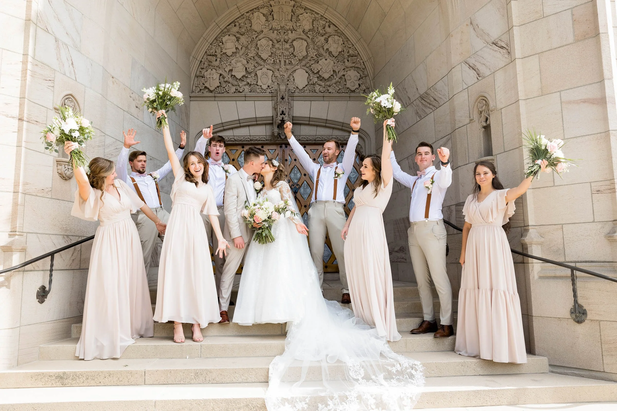 A wedding celebration with a bride and groom kissing surrounded by wedding party members, all celebrating on the steps of a church. The wedding party includes bridesmaids and groomsmen holding bouquets and raising their fists in celebration.
