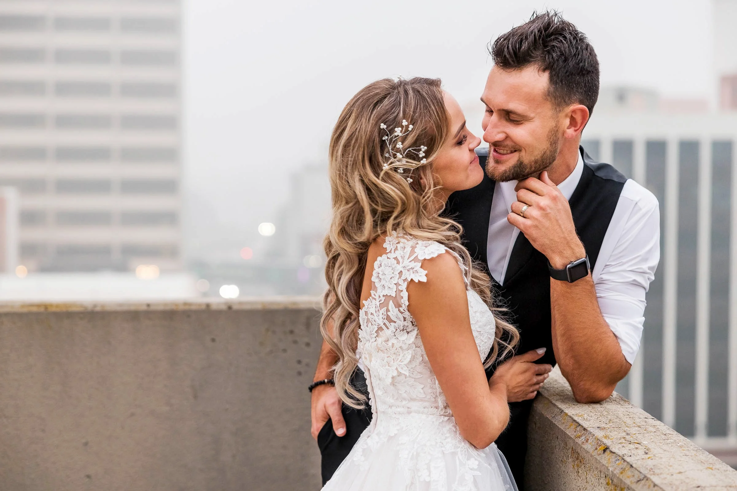 A bride and groom on a rooftop, leaning close and smiling at each other. The bride wears a lace wedding gown with floral details, and has wavy hair decorated with a hairpin. The groom wears a black vest over a white shirt, with a smartwatch and weddi