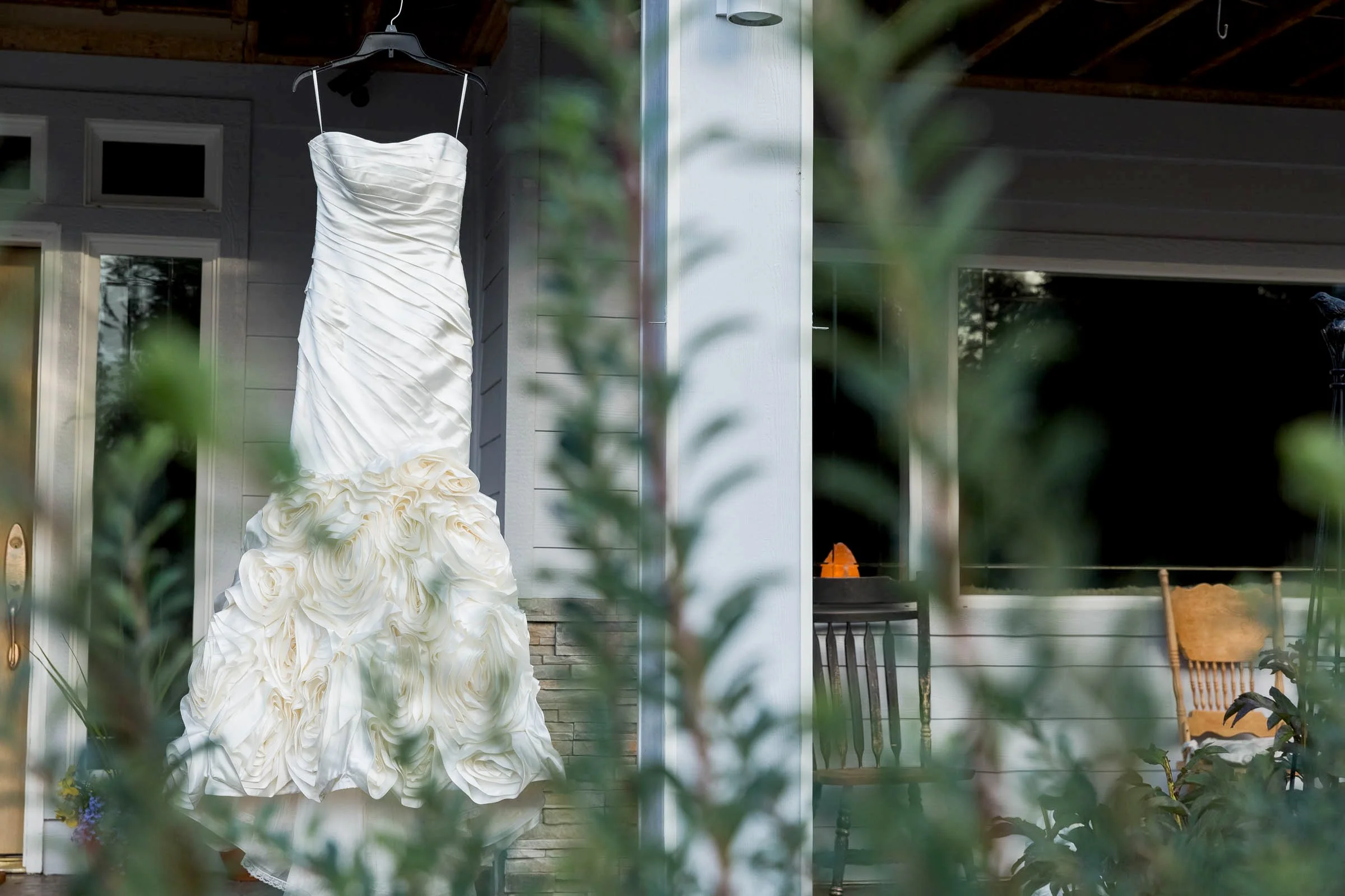 A white wedding dress with ruched top and ruffled floral bottom hanging on a black hanger outside a house.