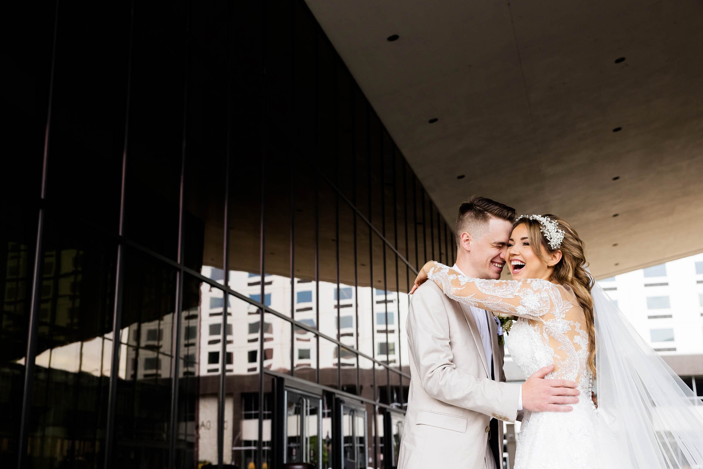 A joyful bride and groom embracing and laughing outside a modern building with reflective glass windows.