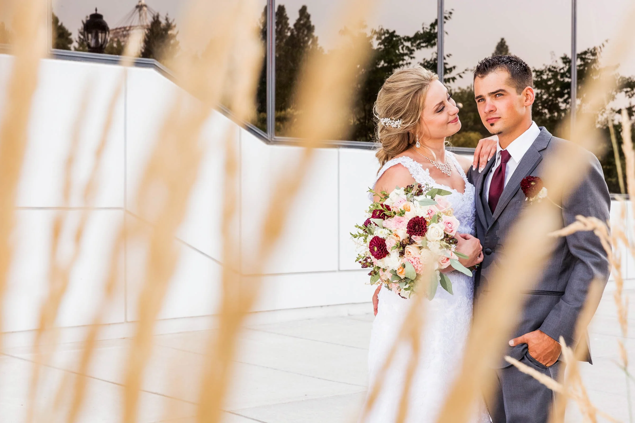 A bride and groom posing outdoors, with the bride holding a bouquet of pink, white, and dark red flowers, while standing in front of a modern white building with large glass windows.