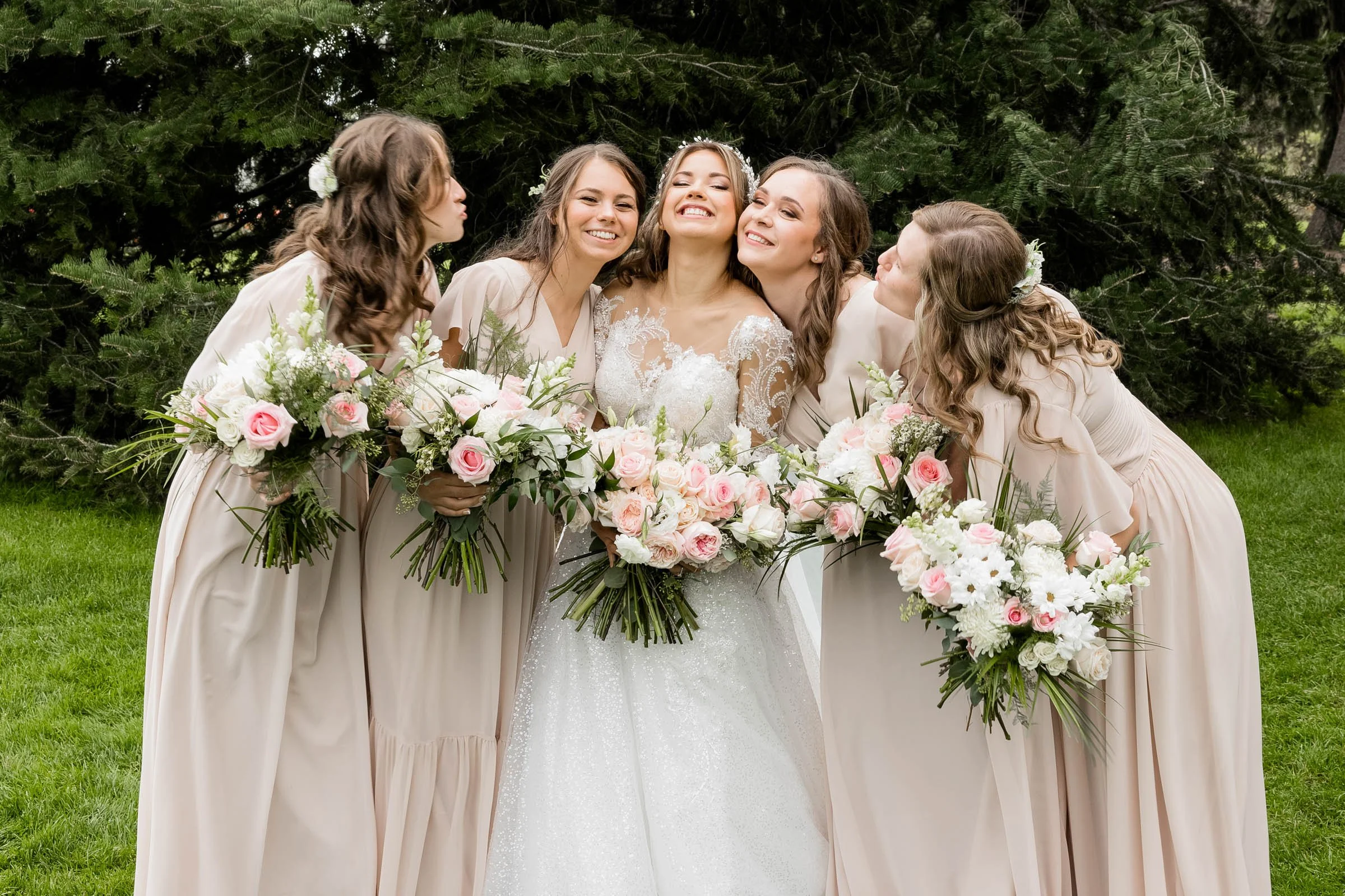 A bride in a wedding gown surrounded by five bridesmaids holding pink and white bouquets, outdoors with green grass and trees in the background.