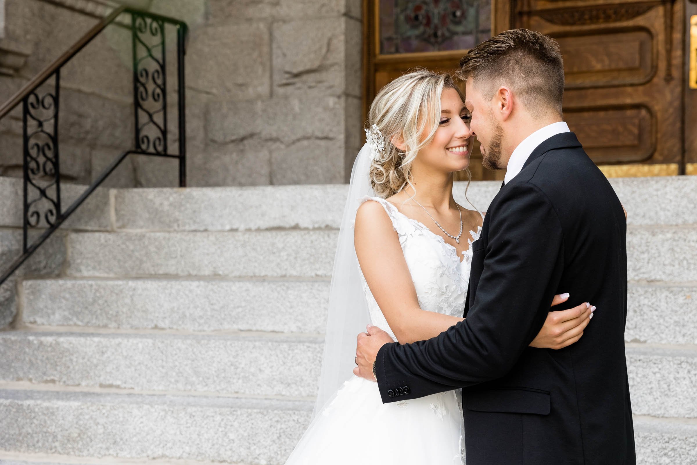 A bride and groom sharing an intimate moment, smiling with eyes closed, in front of stone steps and a building entrance.