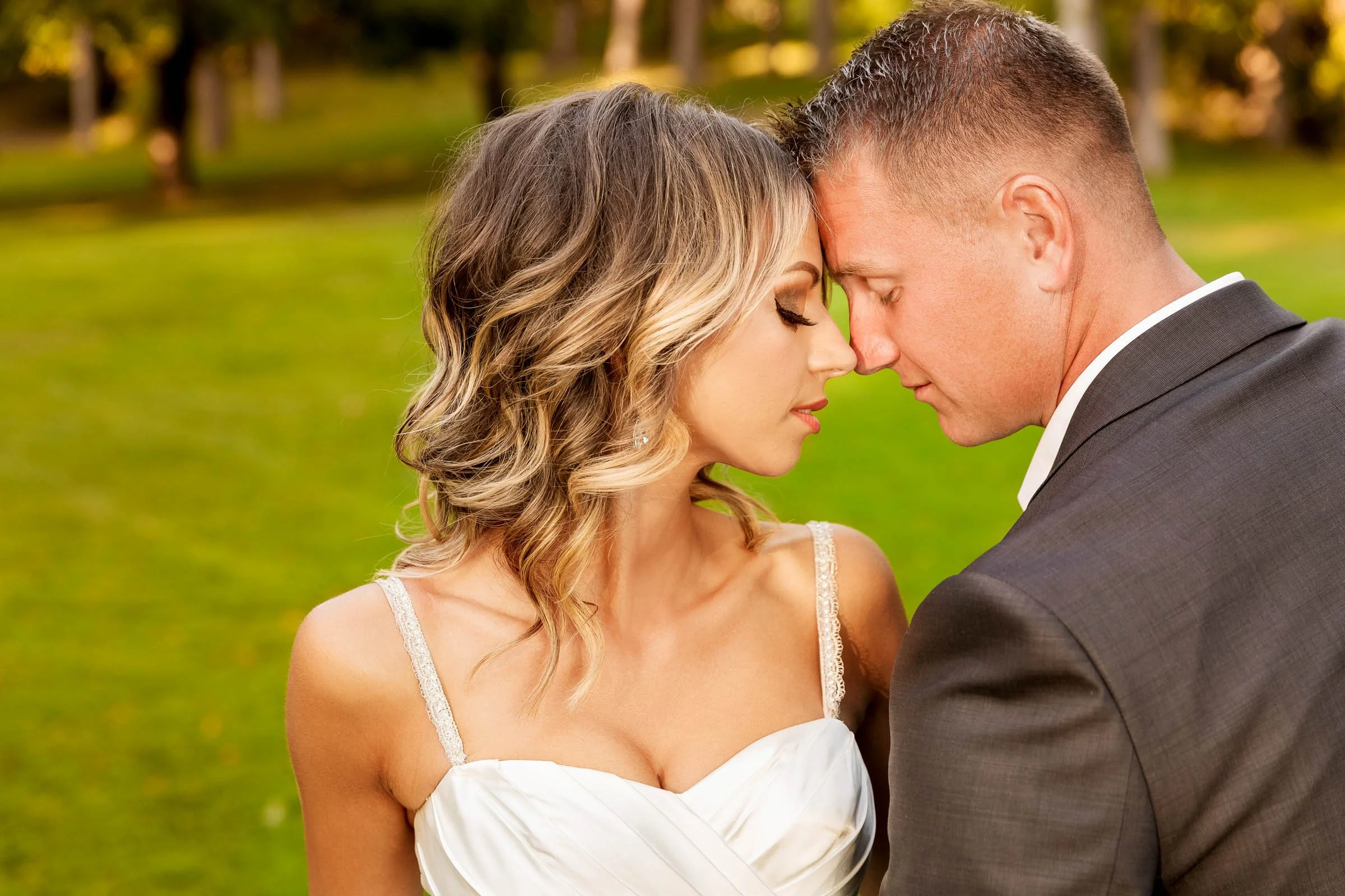 A couple in wedding attire share an intimate moment outdoors, with their foreheads touching and eyes closed, surrounded by green grass and trees.