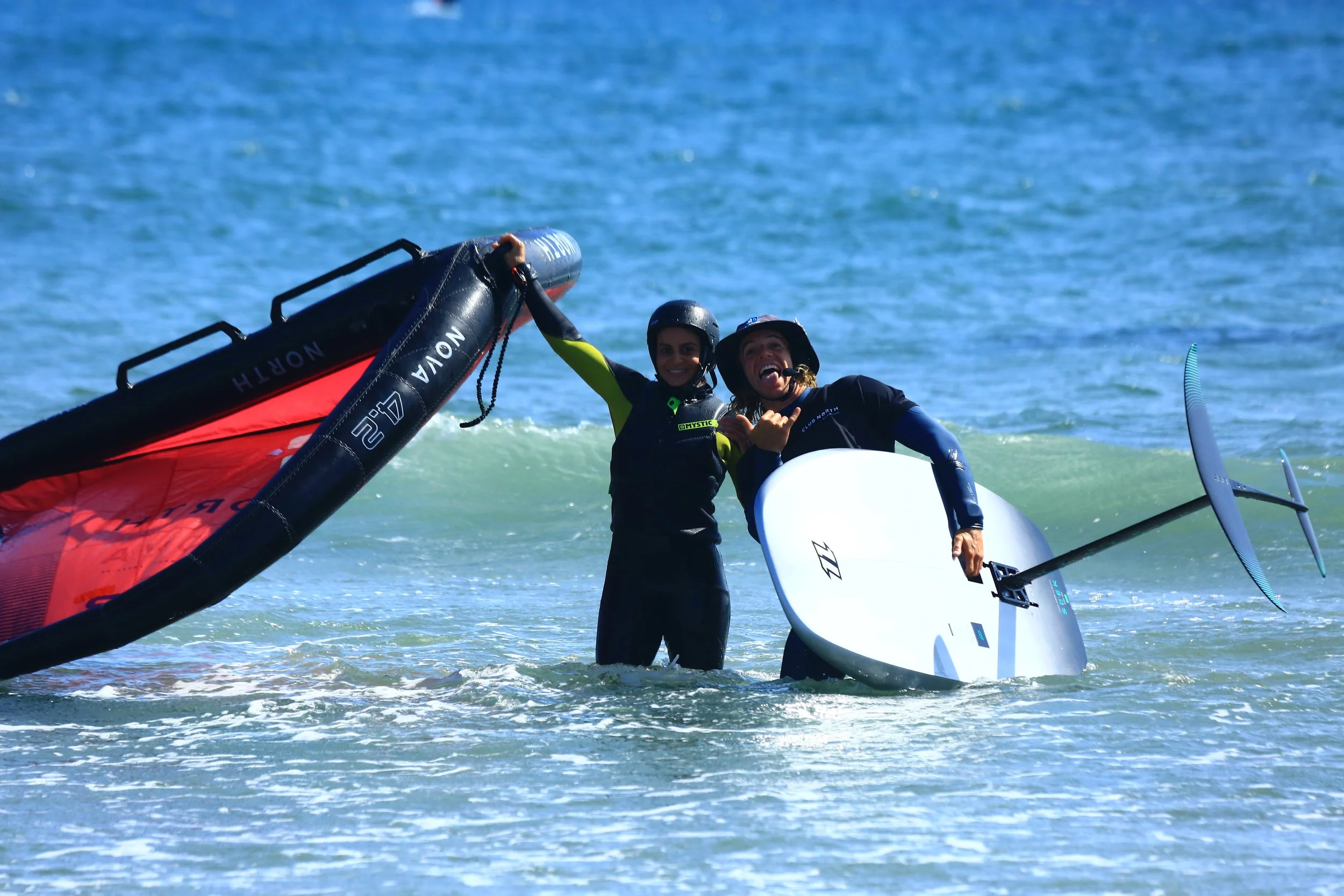 Two people in wetsuits and helmets standing in the ocean with wakesurfing equipment, celebrating after a successful session.