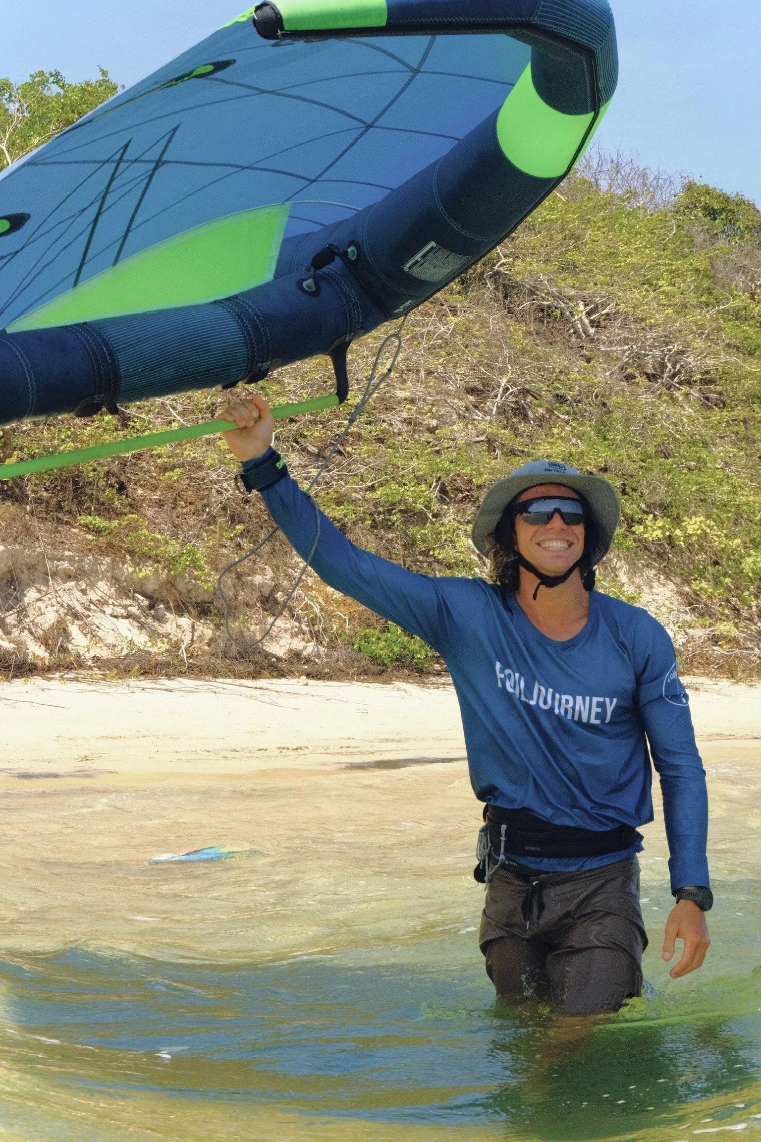 Person standing in shallow water on a beach, holding a kite with a big sail. The person is smiling, wearing sunglasses, a wide-brimmed hat, a blue long-sleeve shirt, and dark shorts.