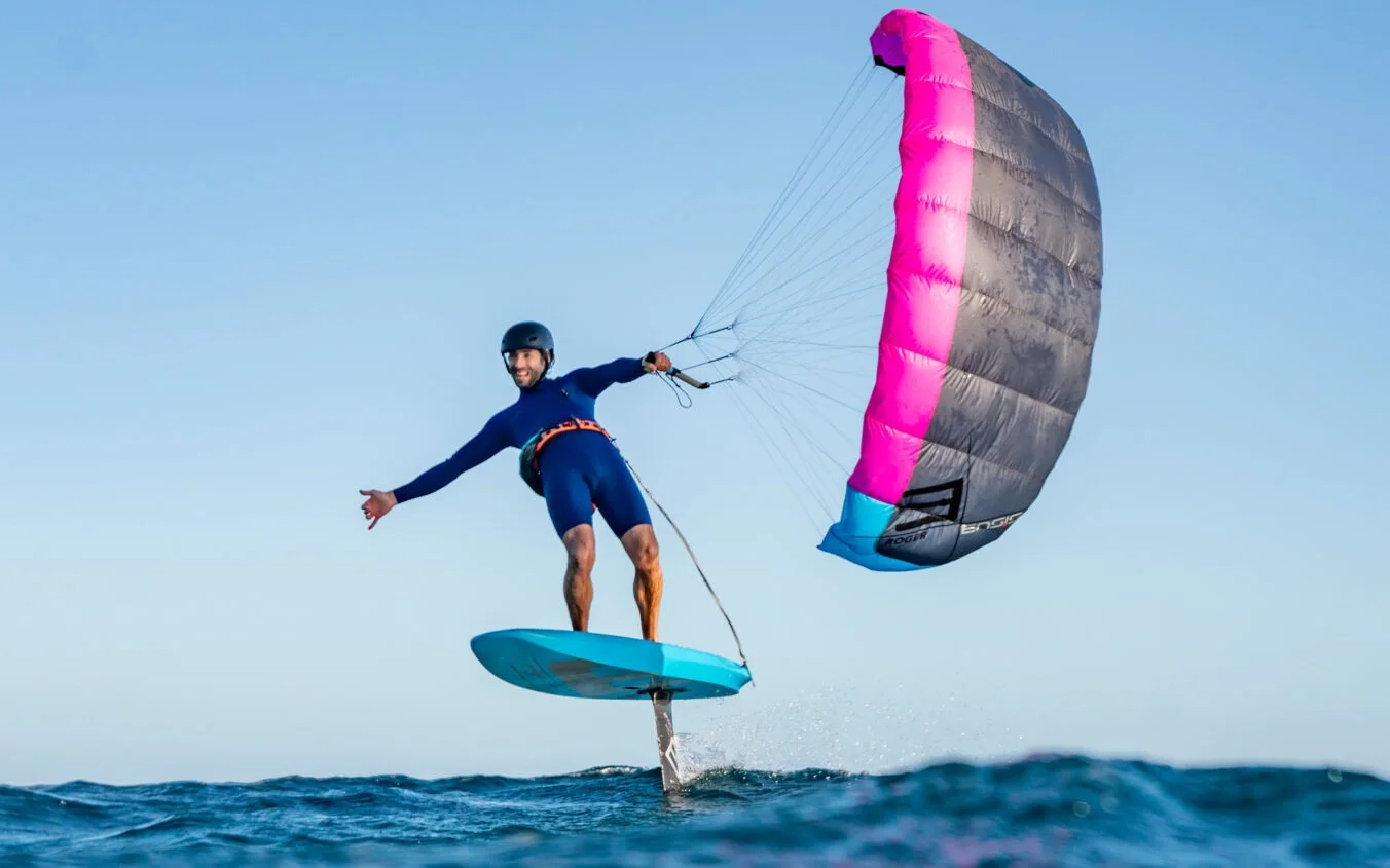 Man parasailing over water on a hydrofoil with a pink and gray parachute, wearing a helmet and wetsuit.