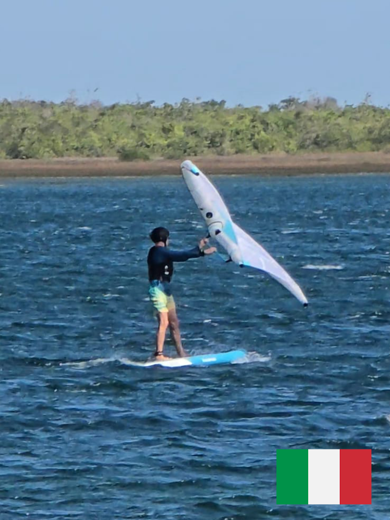 A man with gray hair wearing a blue t-shirt and beige pants sitting on a beige upholstered bench against a plain wall, casting a shadow.