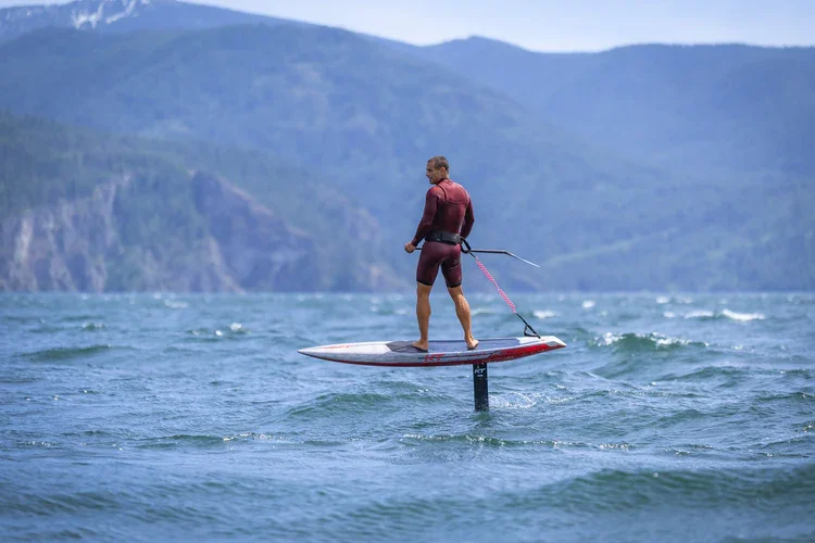A man riding a paddleboard equipped with a motor on a lake, with mountainous terrain in the background.