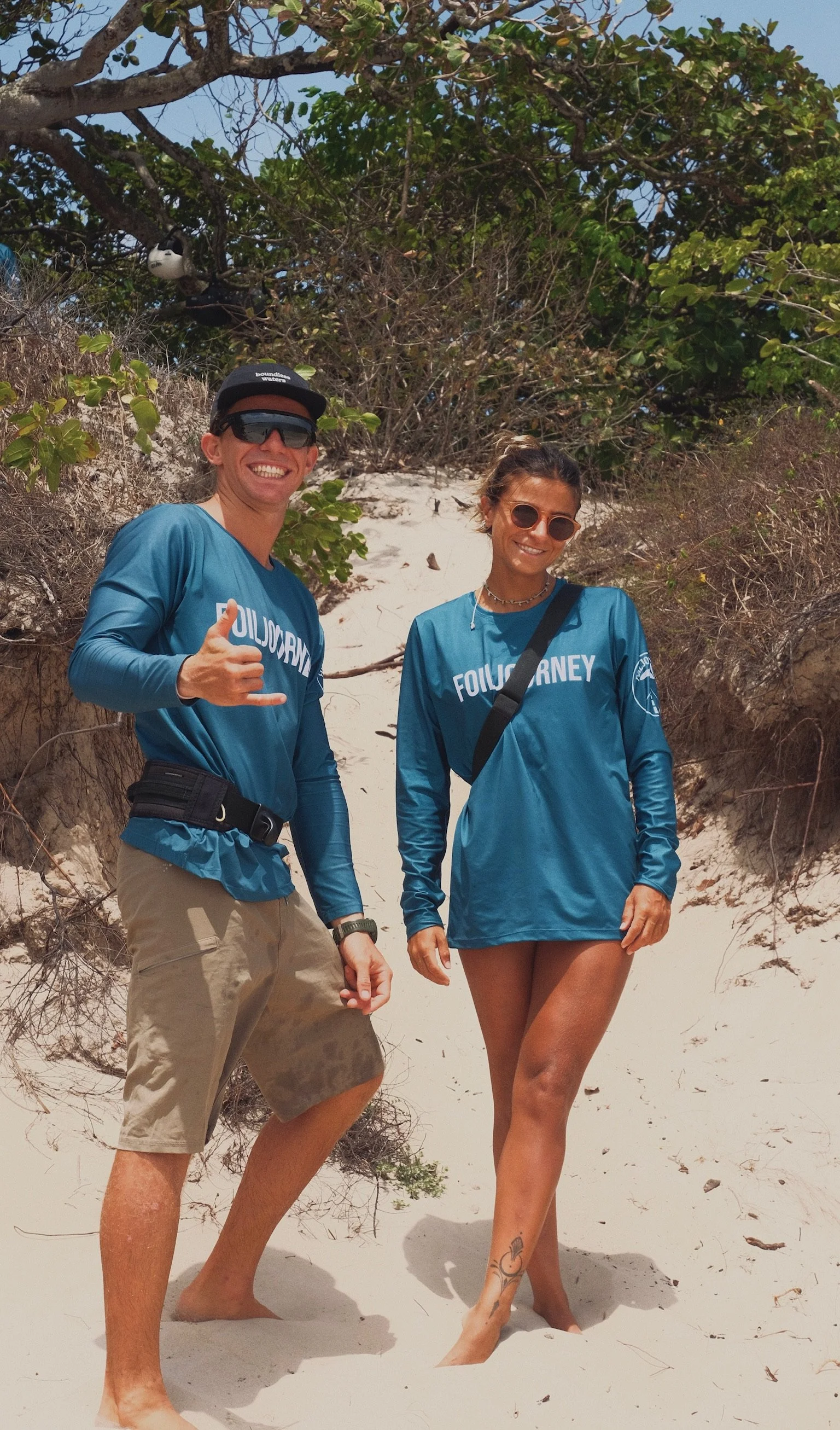 Two people standing on a sandy beach with greenery behind them, wearing matching blue long-sleeve shirts that say 'Follow Your'.