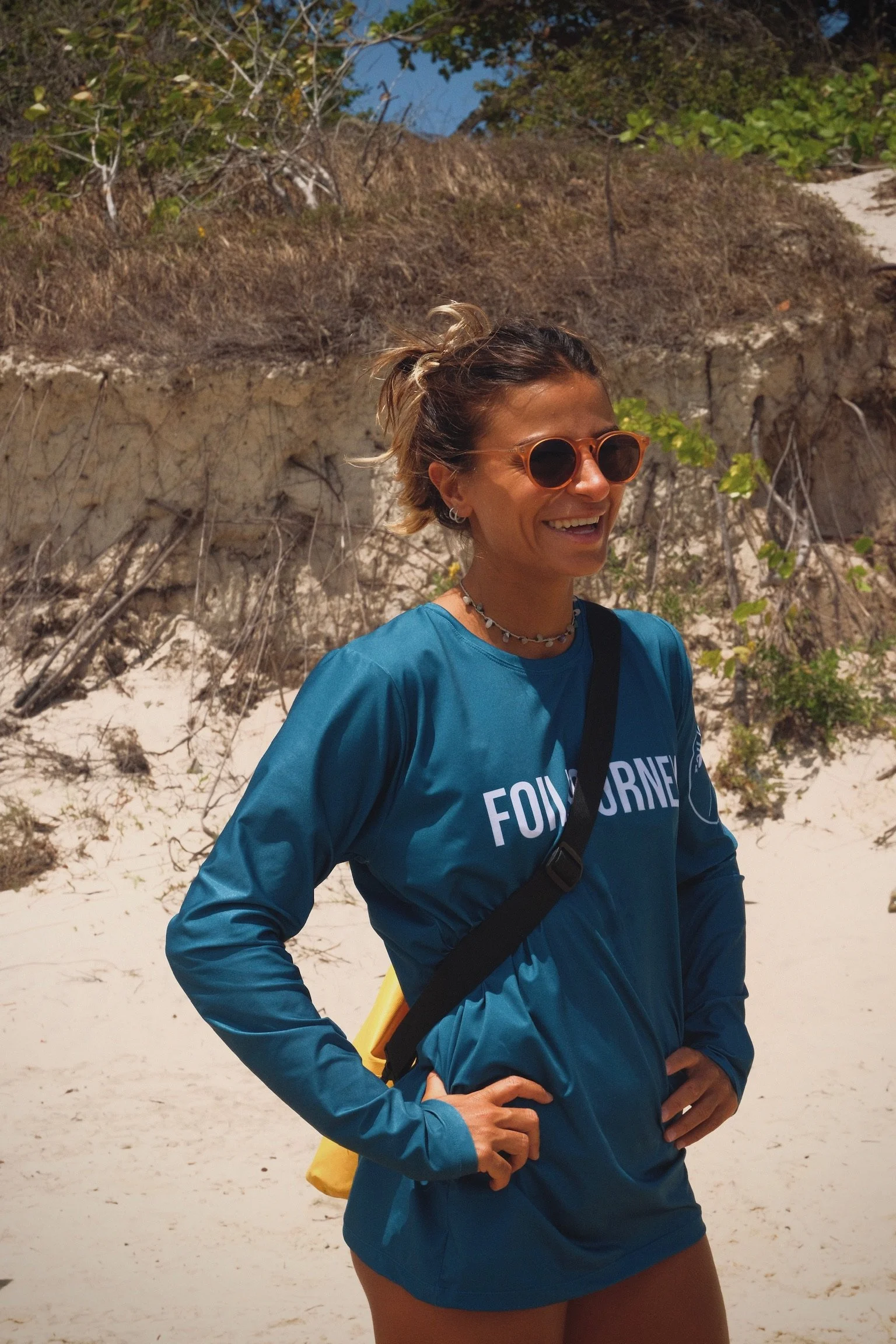 A woman with sunglasses and a blue long-sleeve shirt smiling on a sandy beach.