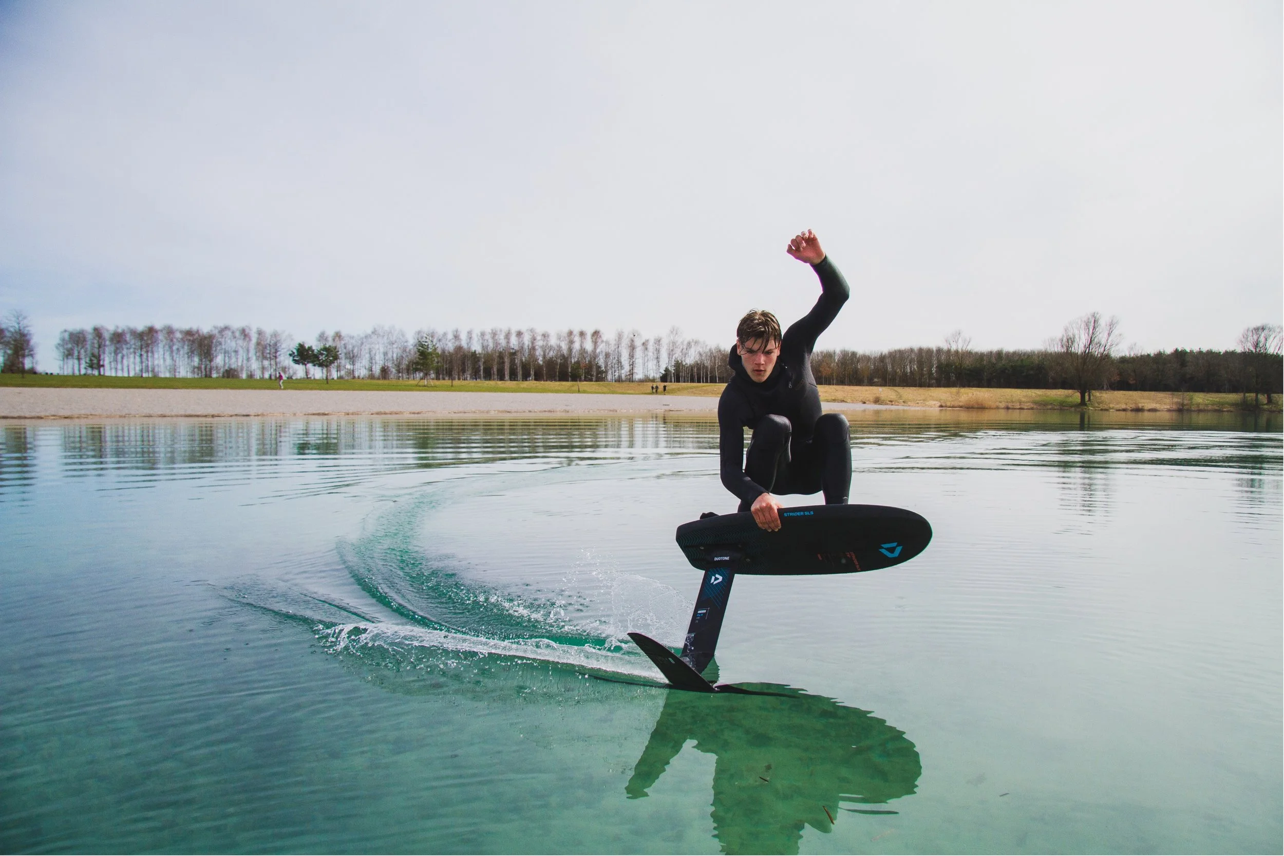 Person in black wetsuit riding a jet surfboard on a calm body of water on a cloudy day.