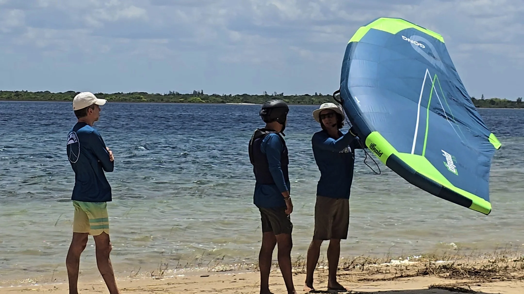 Three men standing on a sandy beach near the water, one holding a blue and green parasail sail, with a distant shoreline in the background.