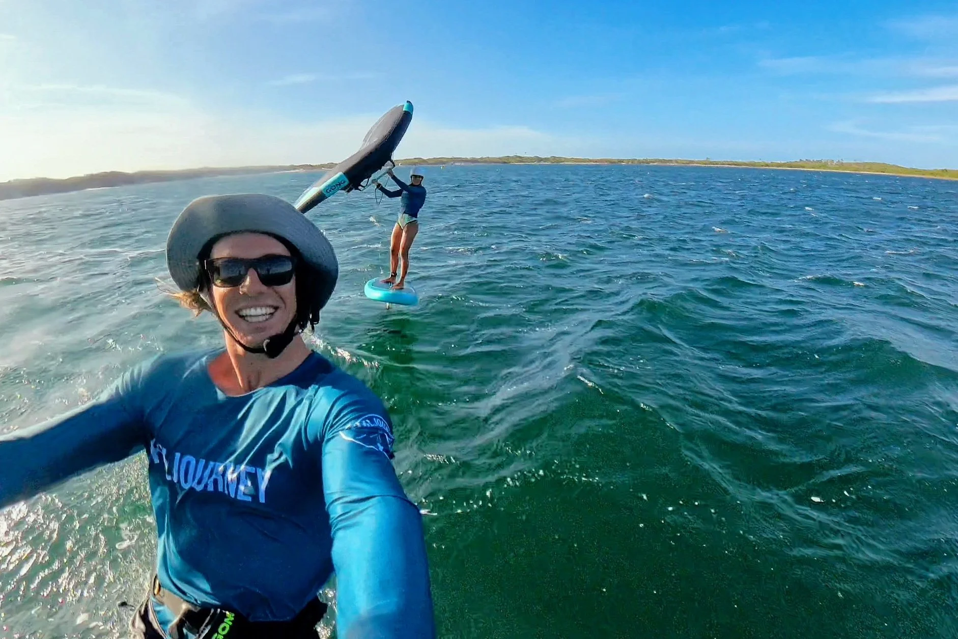 Two women on the water, one taking a selfie with a GoPro and the other riding an efoil board in the background