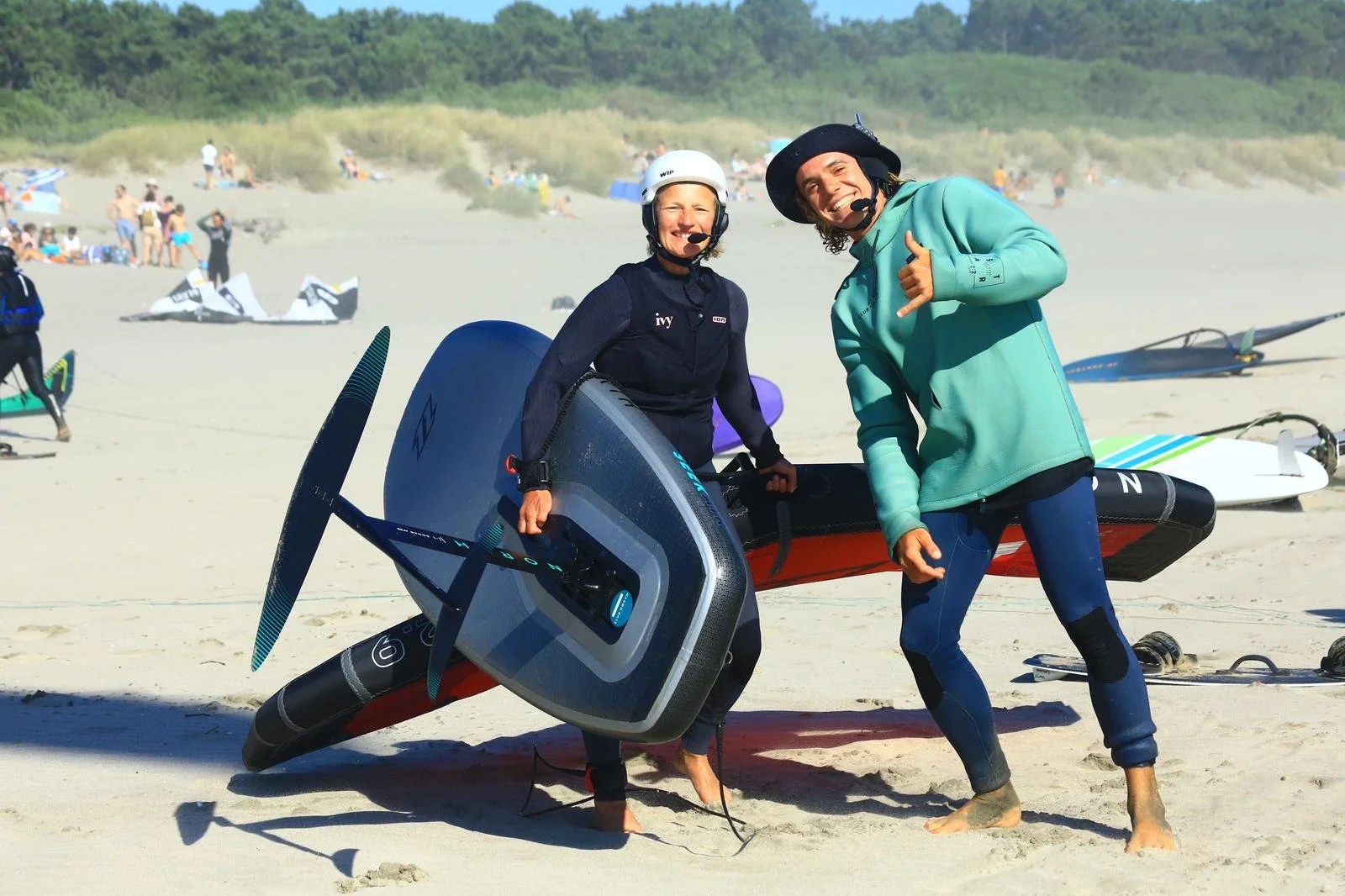 Two people standing on a beach with paddleboards, smiling at the camera, one holding a paddleboard, both wearing wetsuits and helmets, with other beachgoers and paddleboards in the background.