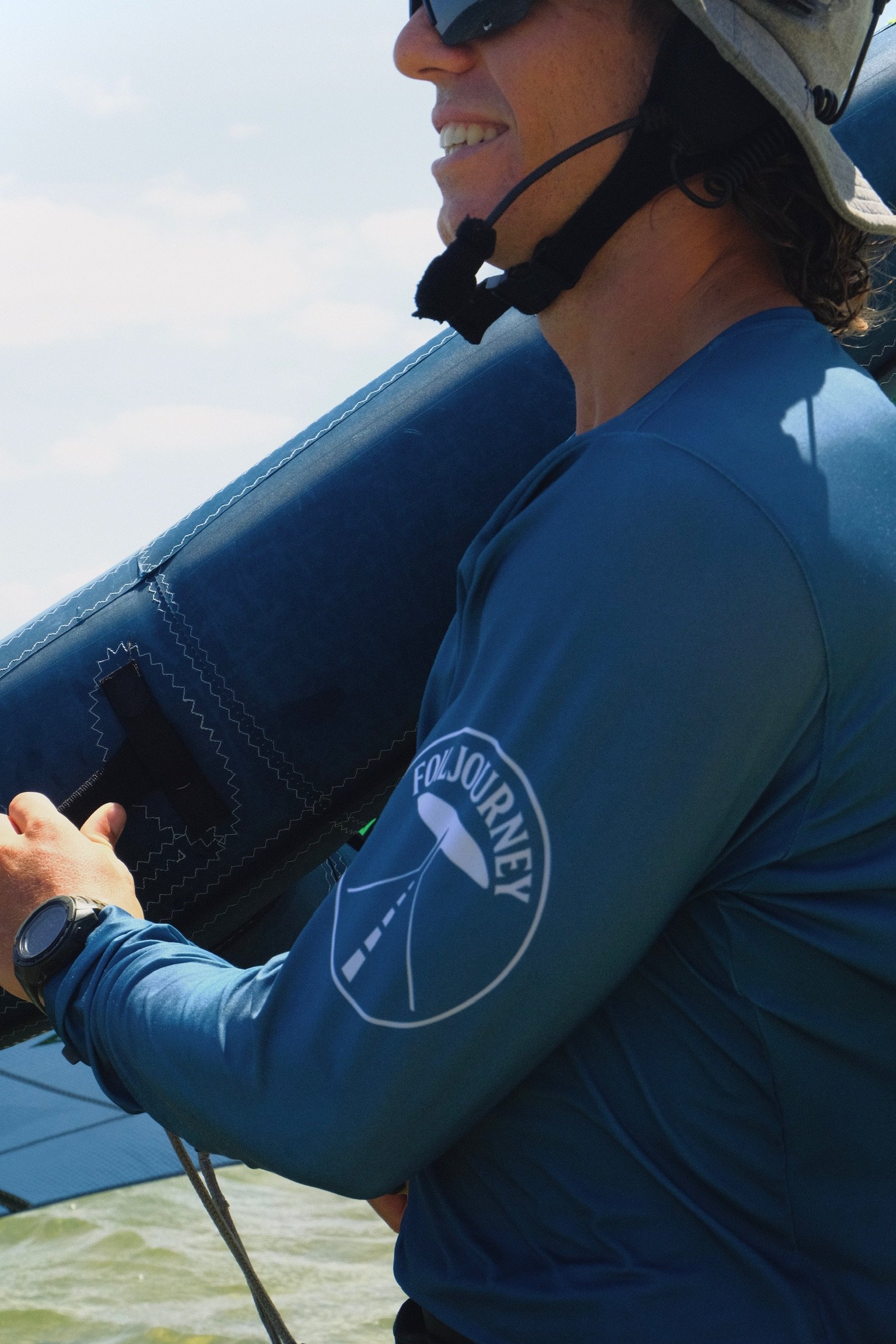 A person wearing a helmet and sunglasses, holding a stand-up paddleboard, and wearing a blue long sleeve shirt with a 'Follow Journeys' logo on the sleeve, standing near the water.