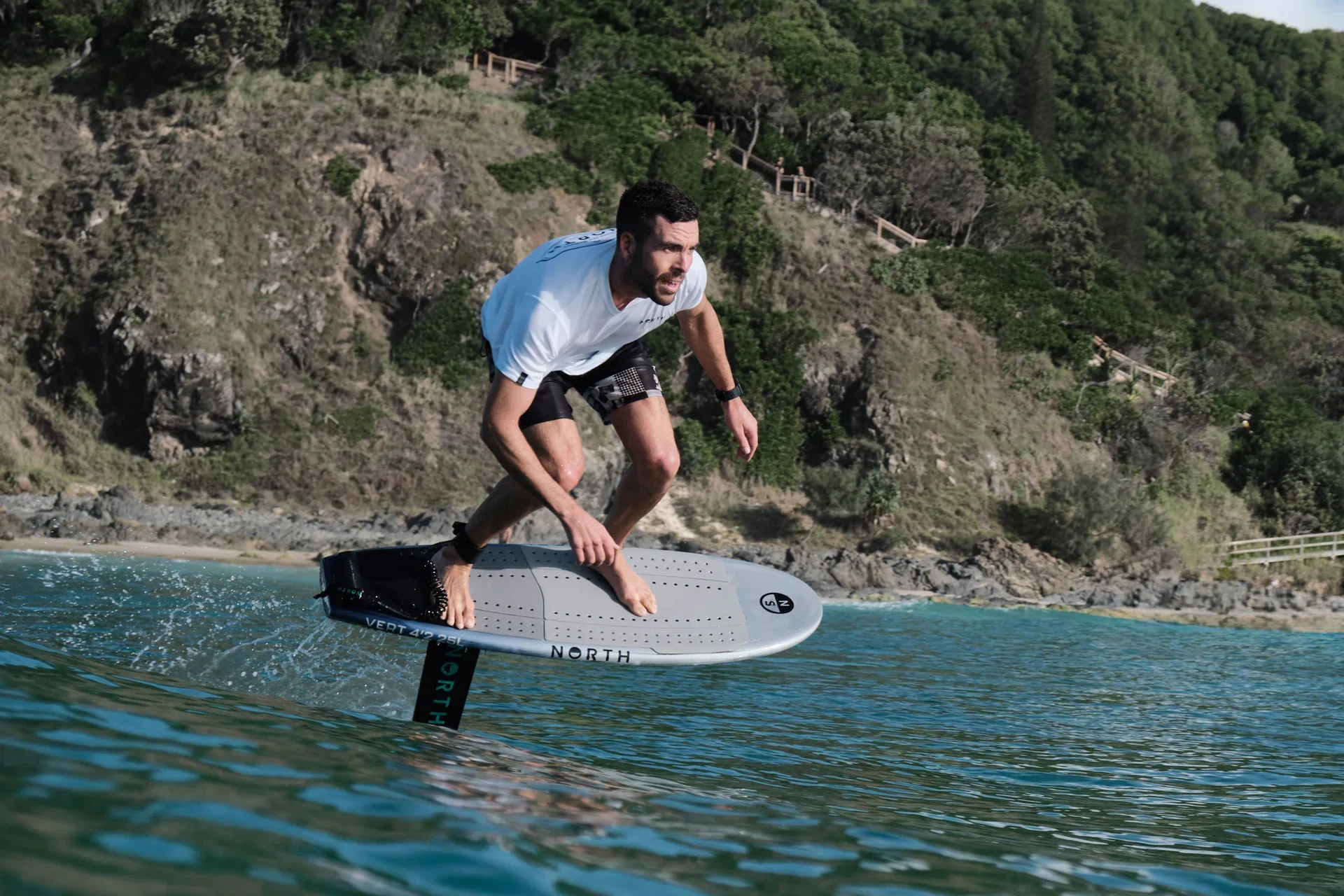 Man surfing on a hydrofoil in the water near a shoreline with hills and trees.
