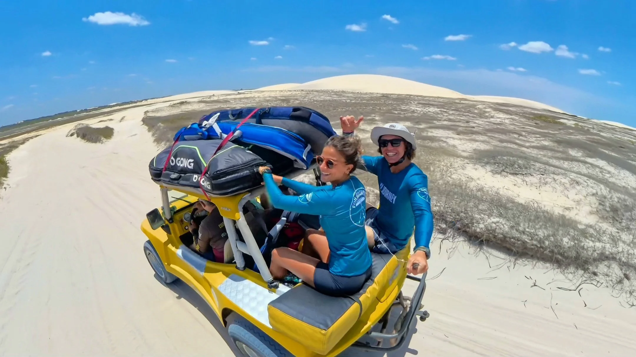 Two women in blue shirts smiling and riding in a yellow off-road vehicle through a sandy desert landscape with sand dunes and a blue sky with clouds.