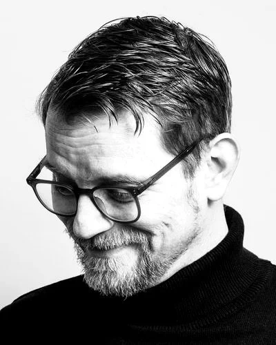 Schwarz-weiß Portrait von Leiv mit Brille und kurzen, gepflegten Haaren, lächelnd mit Blick nach unten.