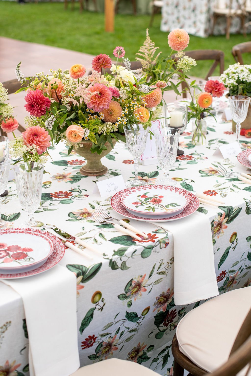 A decorated outdoor table with a floral tablecloth, pink and white plates with floral patterns, clear glassware, and a large pink and white flower centerpiece.