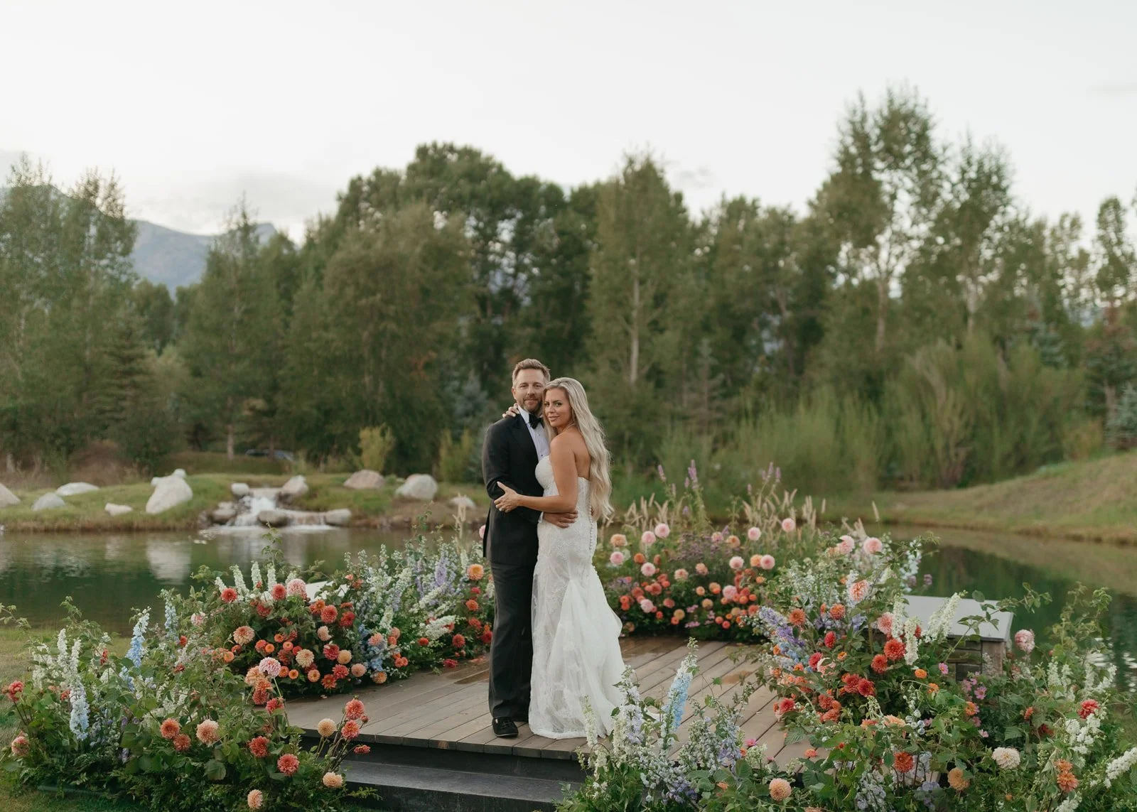 A bride and groom in wedding attire standing on a platform surrounded by colorful flowers, next to a body of water with trees and mountains in the background.