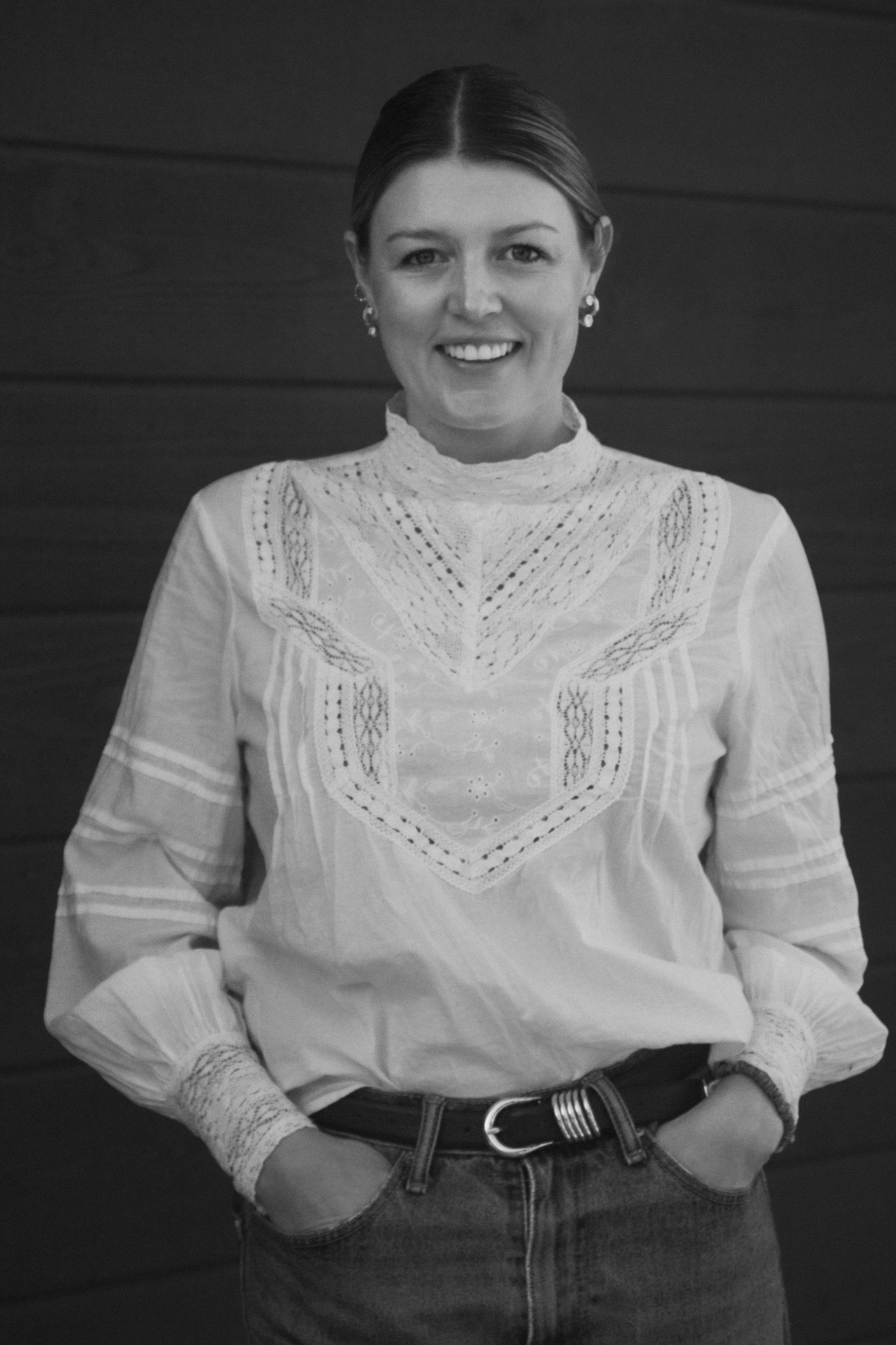 Black and white photo of a woman with short straight hair, smiling, wearing a high-neck embroidered blouse, earrings, a belt, and jeans, standing against a wooden background.