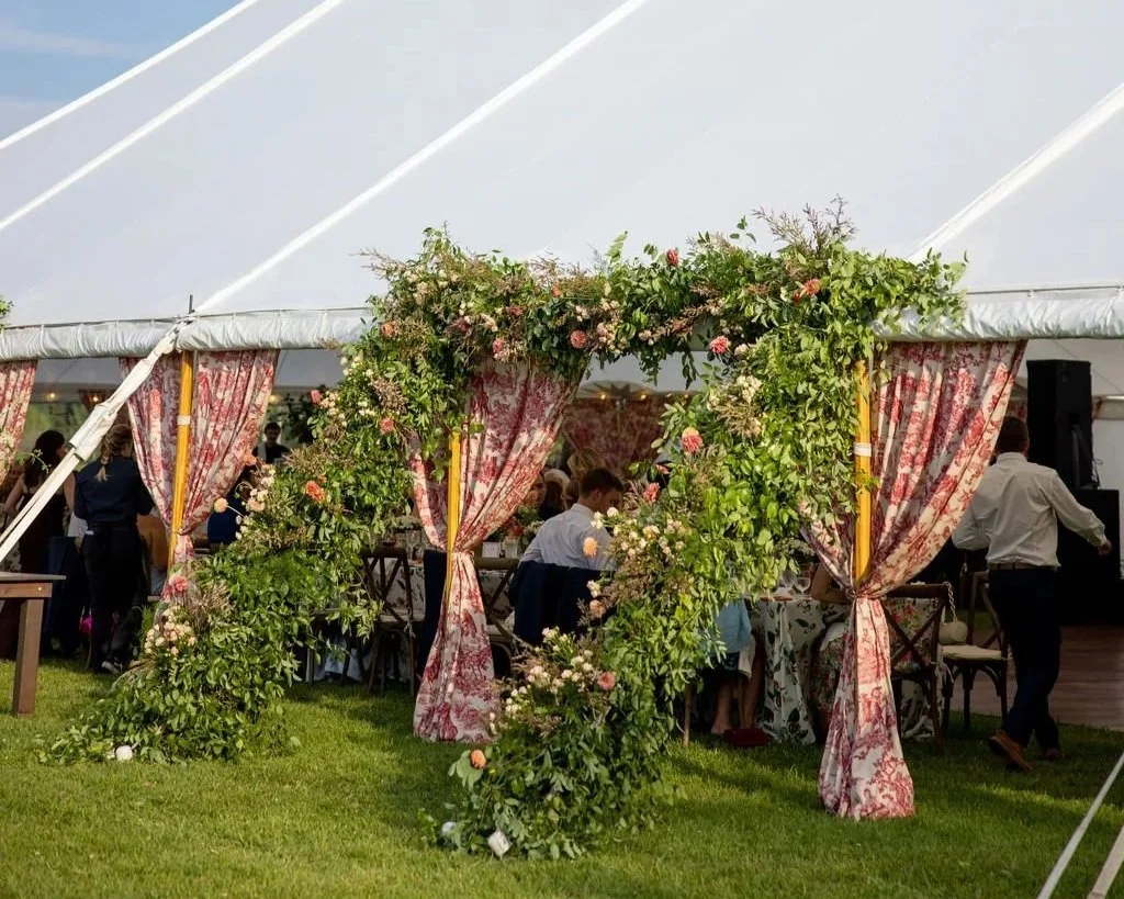 A decorated wedding or event tent with floral archway and red patterned curtains, hosting guests inside on a grassy outdoor setting.