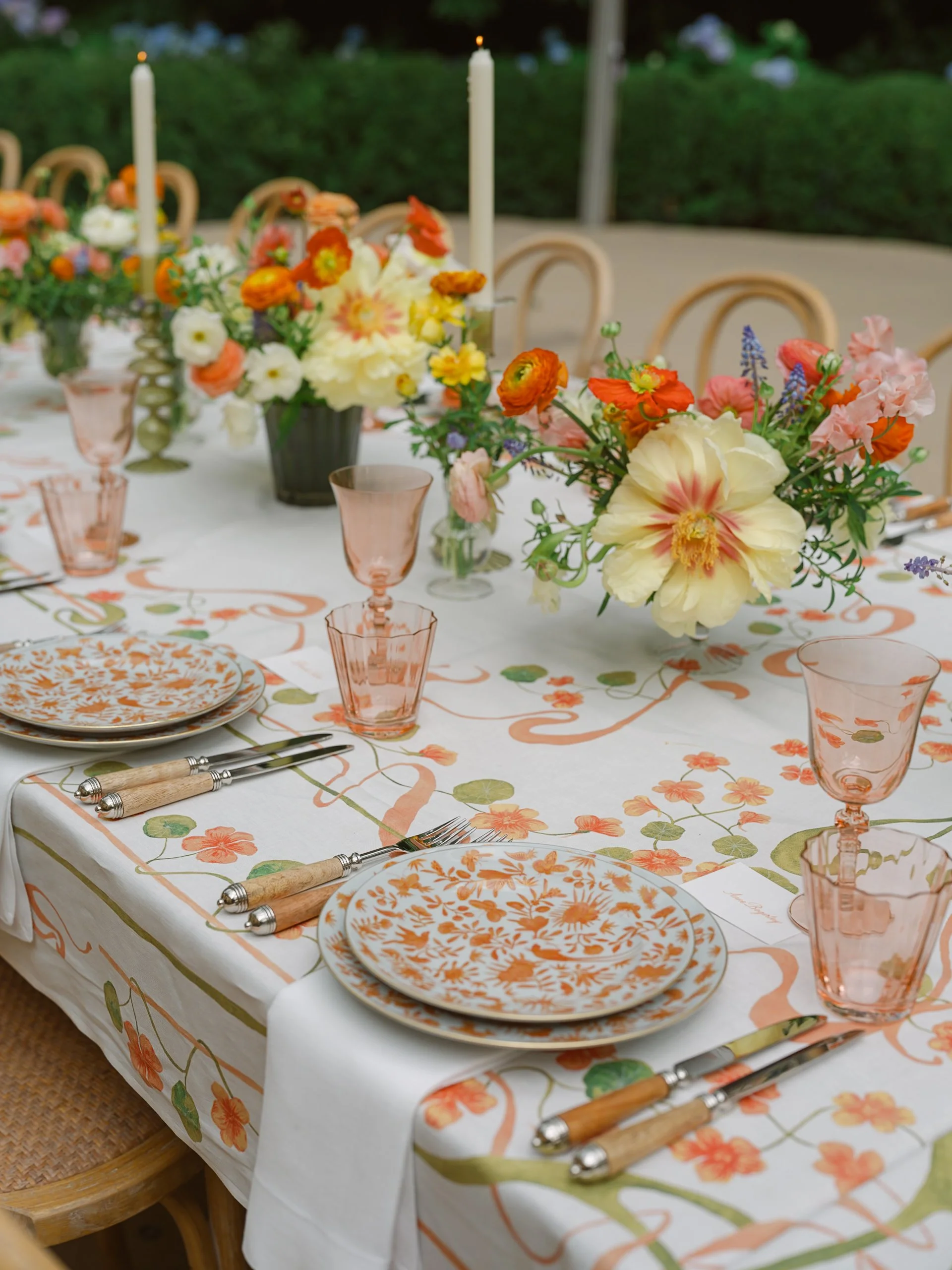 A decorated dining table with floral centerpiece, plates, glasses, and utensils set for a meal, outdoors with wicker chairs around.