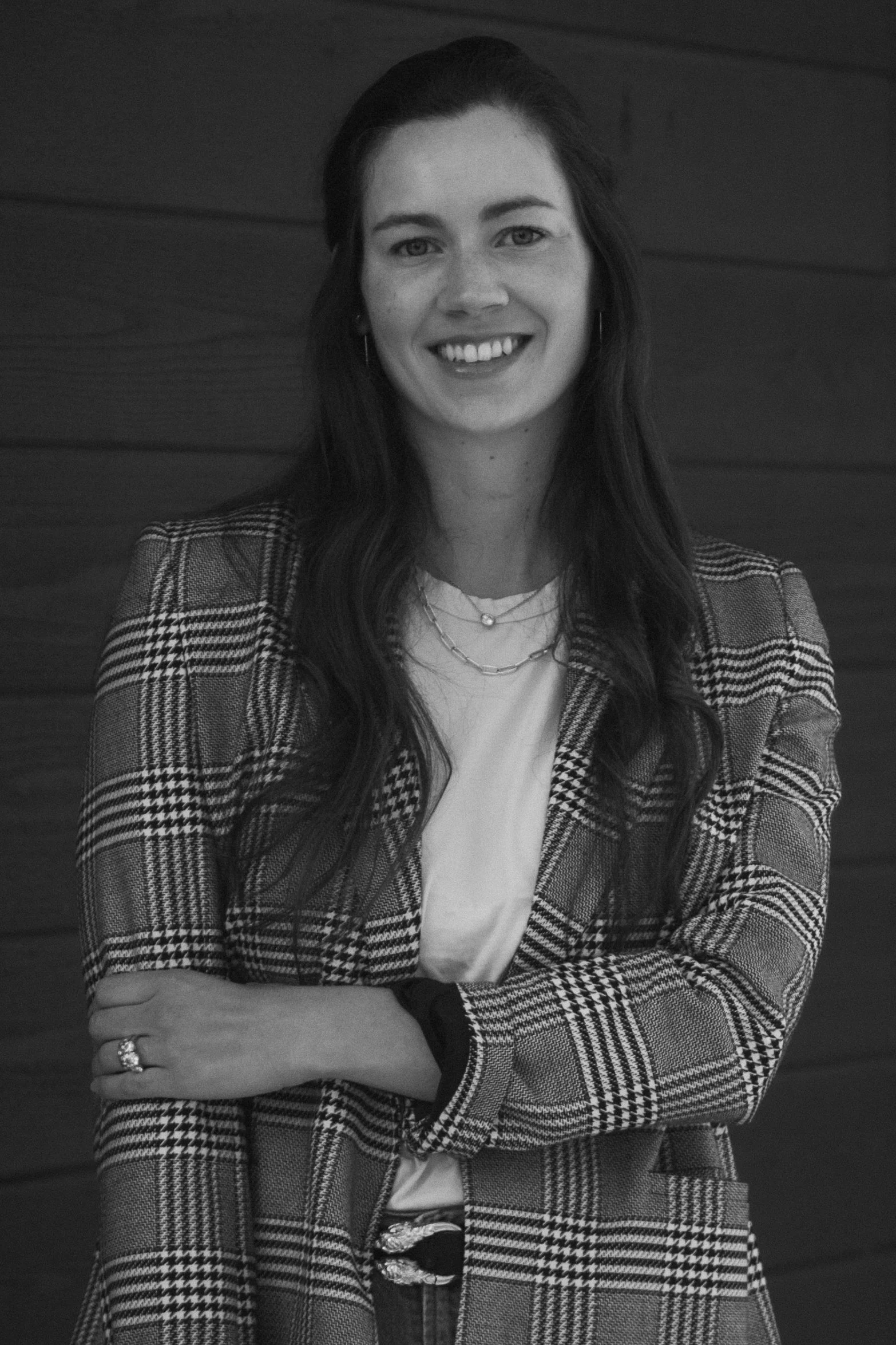 Black and white photo of a smiling woman with long dark hair, wearing a plaid blazer, layered necklaces, and earrings, with her arms crossed, standing against a wood-paneled wall.