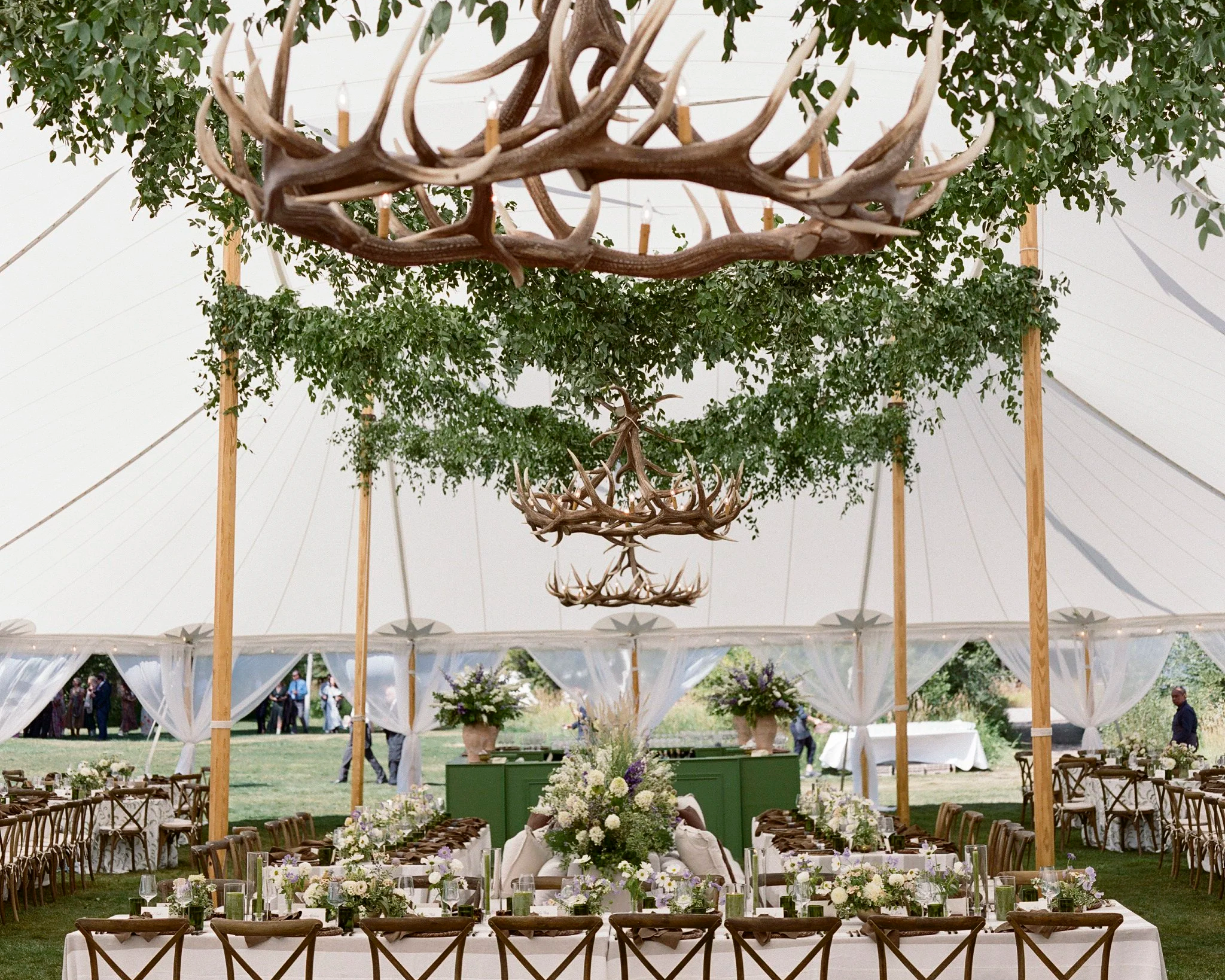 Outdoor event space decorated with chandeliers made of antlers, surrounded by greenery, with tables set with flowers and candles.