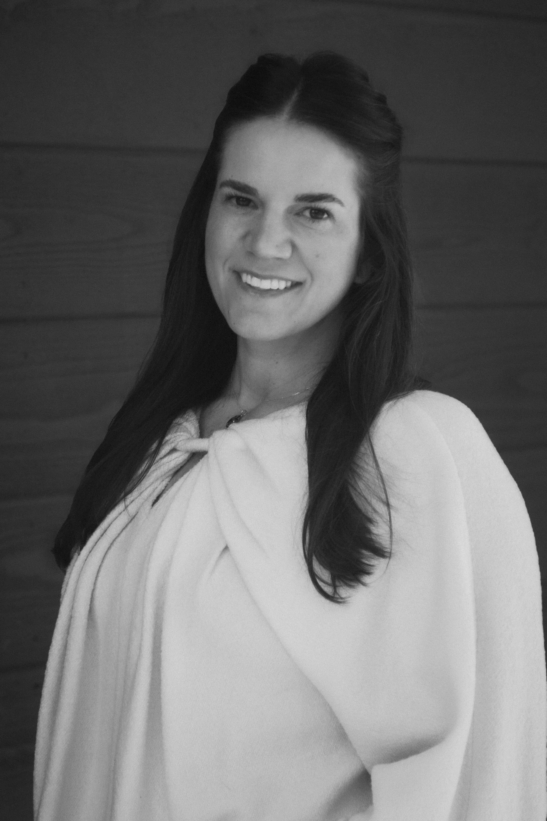 A black and white portrait of a woman with long dark hair, smiling, with a wooden background.