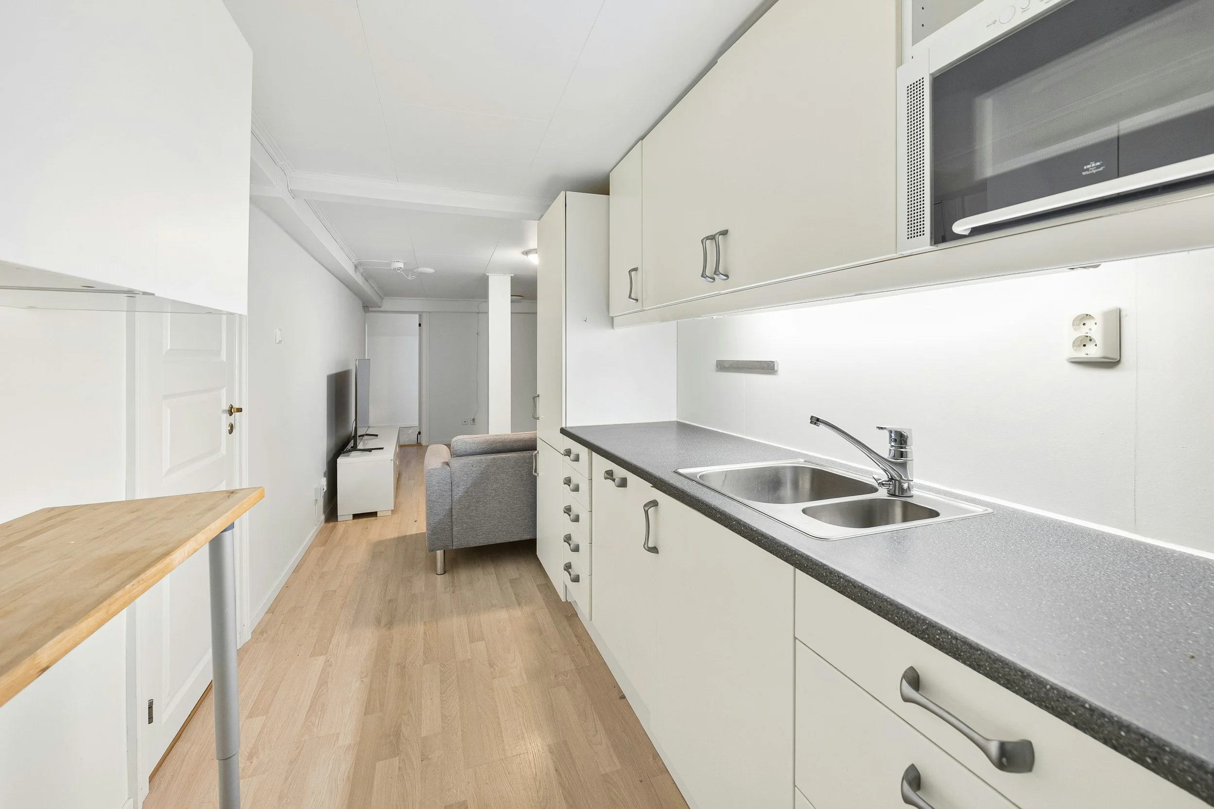 View of a minimalist kitchen and living room with light wood flooring, white walls, white cabinets, a grey countertop, a double sink, and a small living area with a TV and a grey sofa.