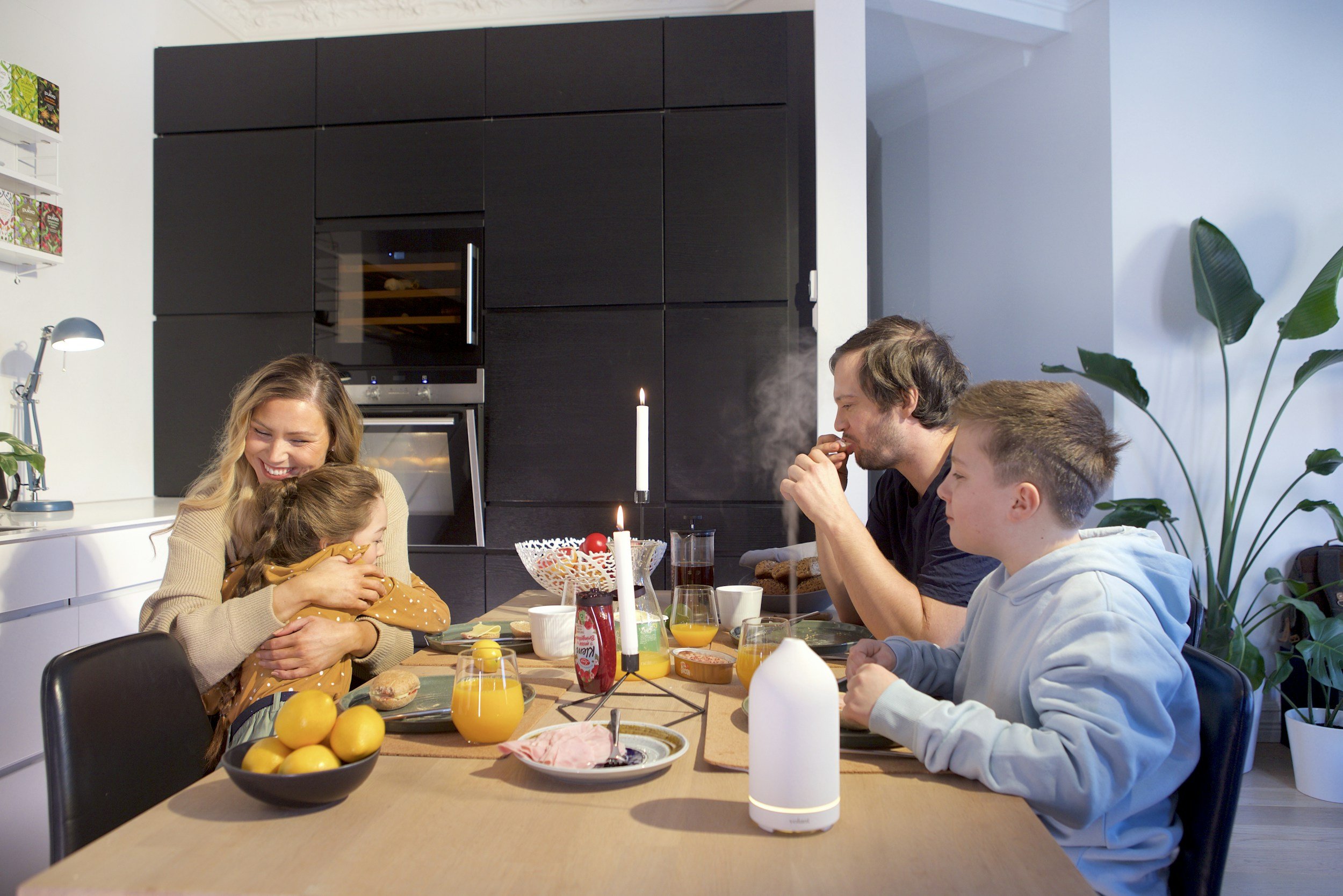 Family enjoying breakfast at the dining table, with a woman hugging a young girl, man eating, and a teenage boy, on a modern kitchen background with plants and breakfast foods.