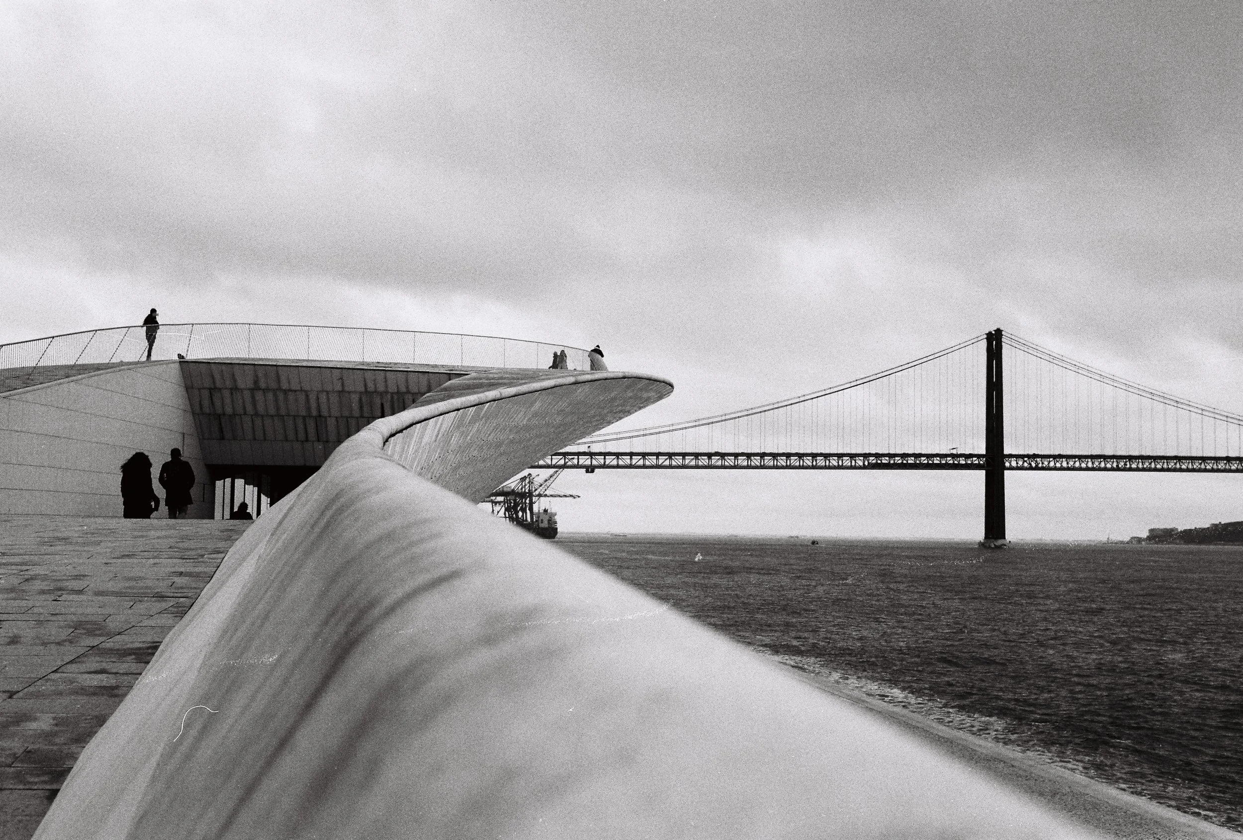 Black and white photo of a modern building with a curved design near a body of water, with a bridge in the background and a cloudy sky. Shot on a Voigtländer Bessa R3A with Cinestill XX film