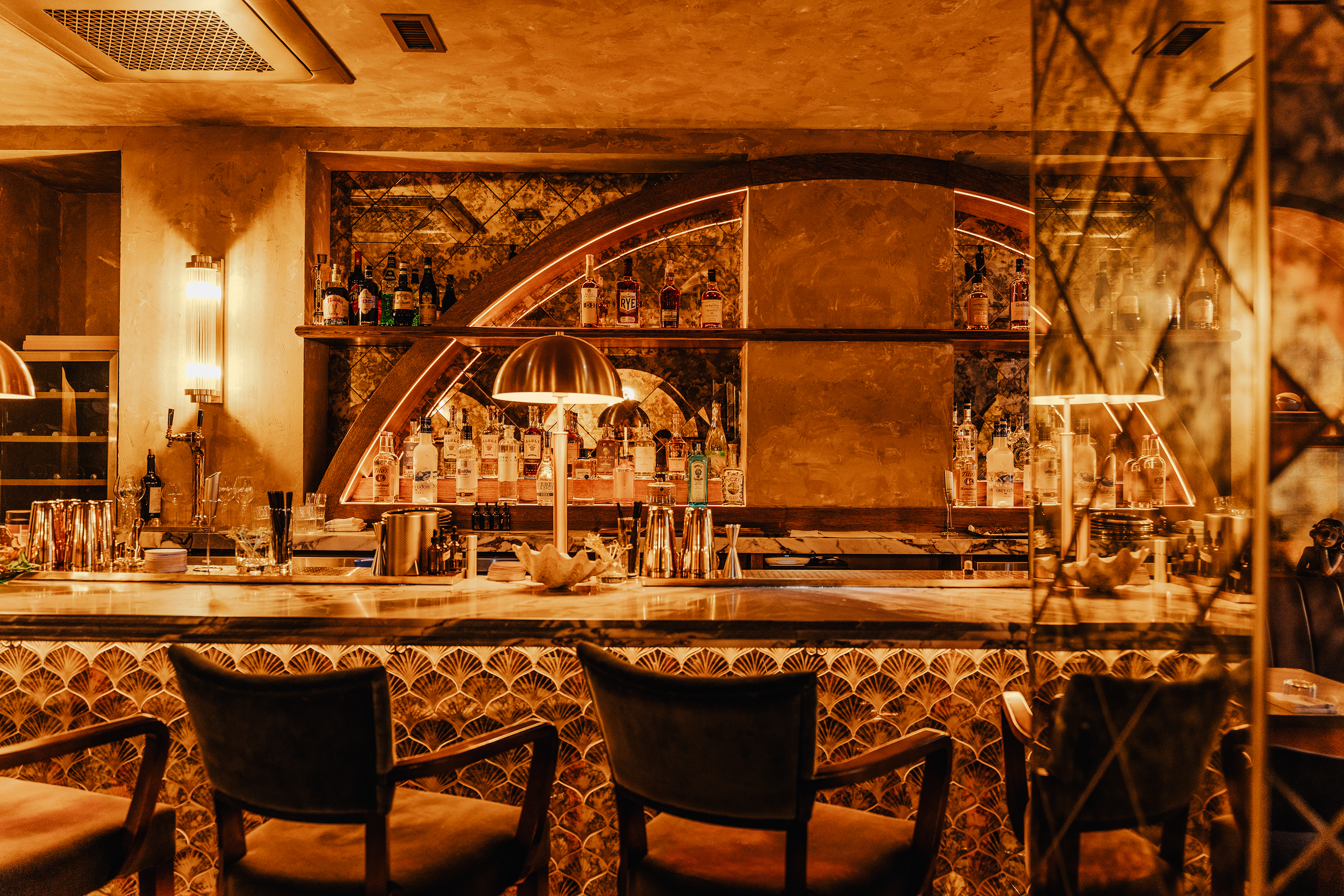 A bar with a marble counter, displaying various bottles of alcohol and barware. Behind the bar, there are shelves with additional bottles, a decorative mirror, and warm lighting creating an inviting atmosphere.