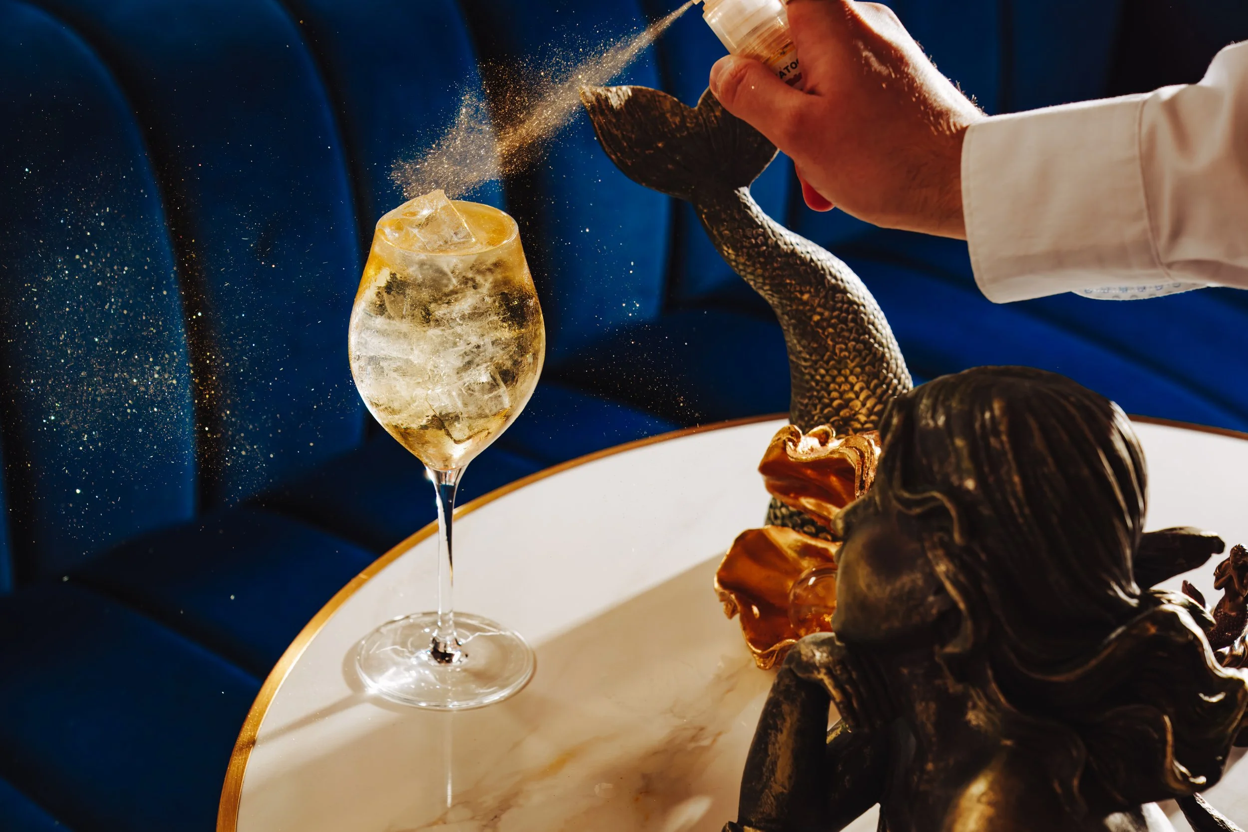 A person holding a salt shaker sprinkling salt onto a wine glass filled with ice, with a blue background and a plate of assorted dried fruits or snacks nearby.