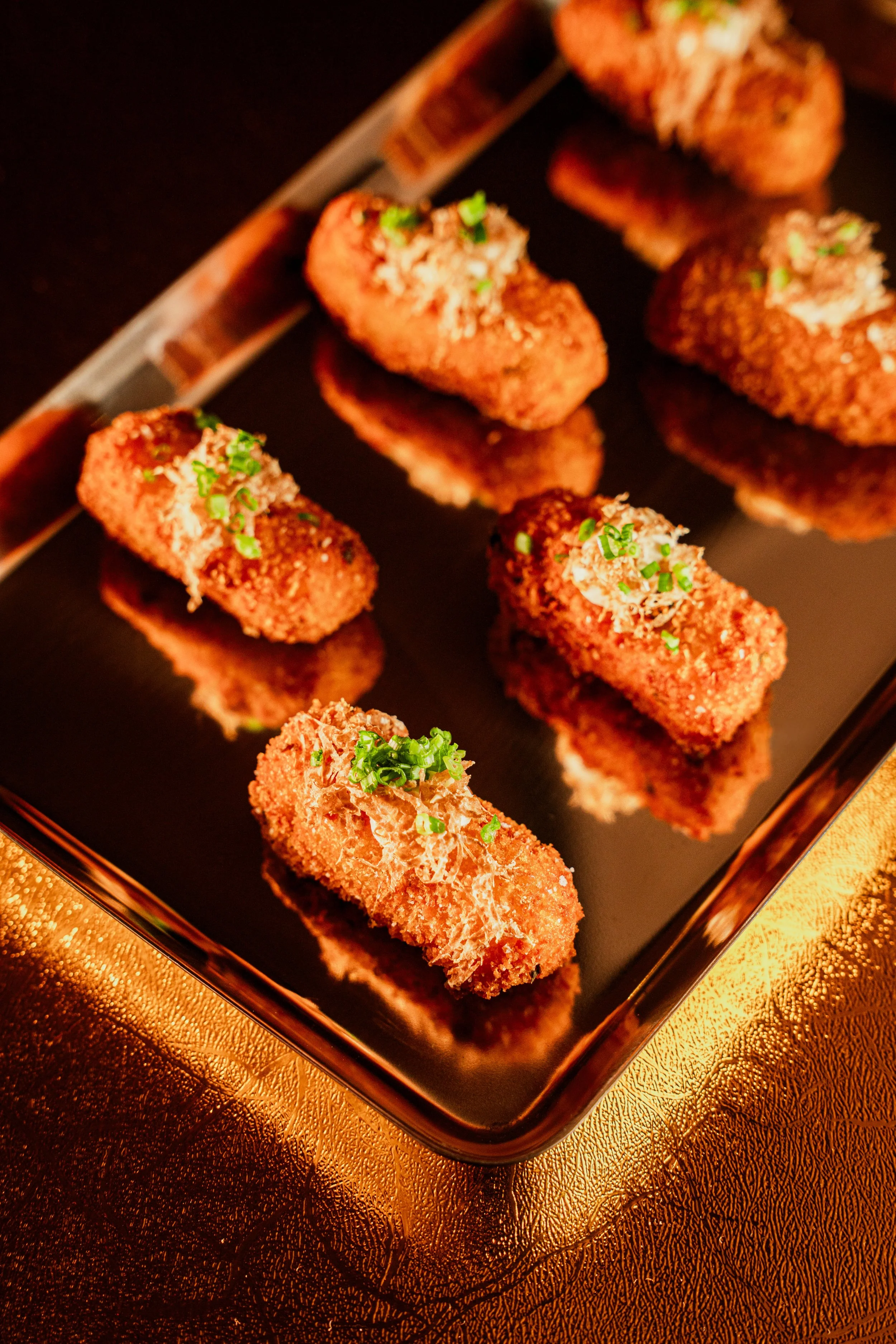 Close-up of breaded and fried croquettes topped with chopped green onions on a black platter, with one croquette cut open showing filling, all on a gold-colored textured surface.