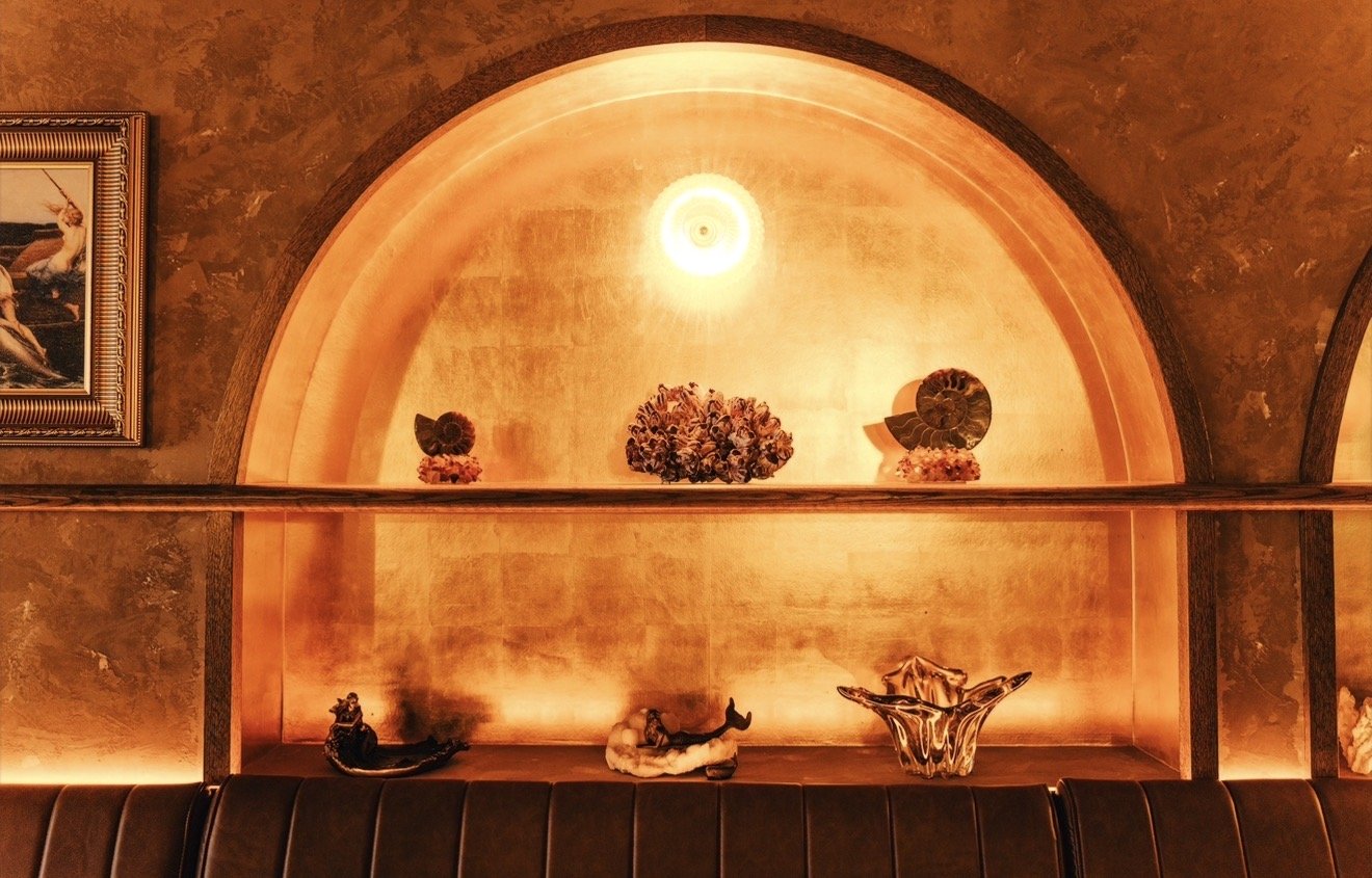 Decorative display with ceramic and floral ornaments against a warm, lit gold back wall, with a framed painting on the left and dark cushioned seating at the bottom.