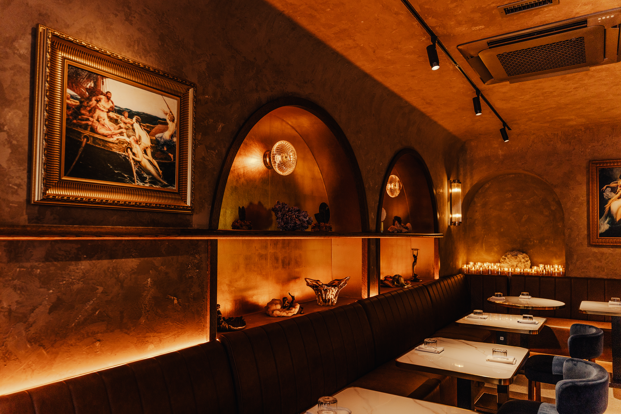 A dimly lit restaurant with warm lighting, dark leather banquettes, and white marble tables set with glassware and napkins. The walls are decorated with framed artwork and decorative sculptures.