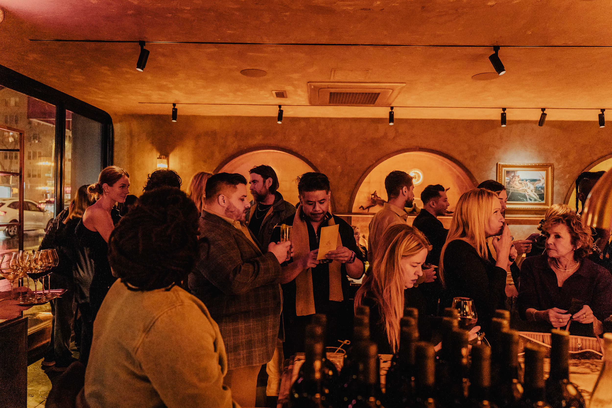 Group of people gathering at a dimly lit restaurant or bar, some are looking at menus, others are engaged in conversation. The decor includes framed art on the walls and track lighting on the ceiling.