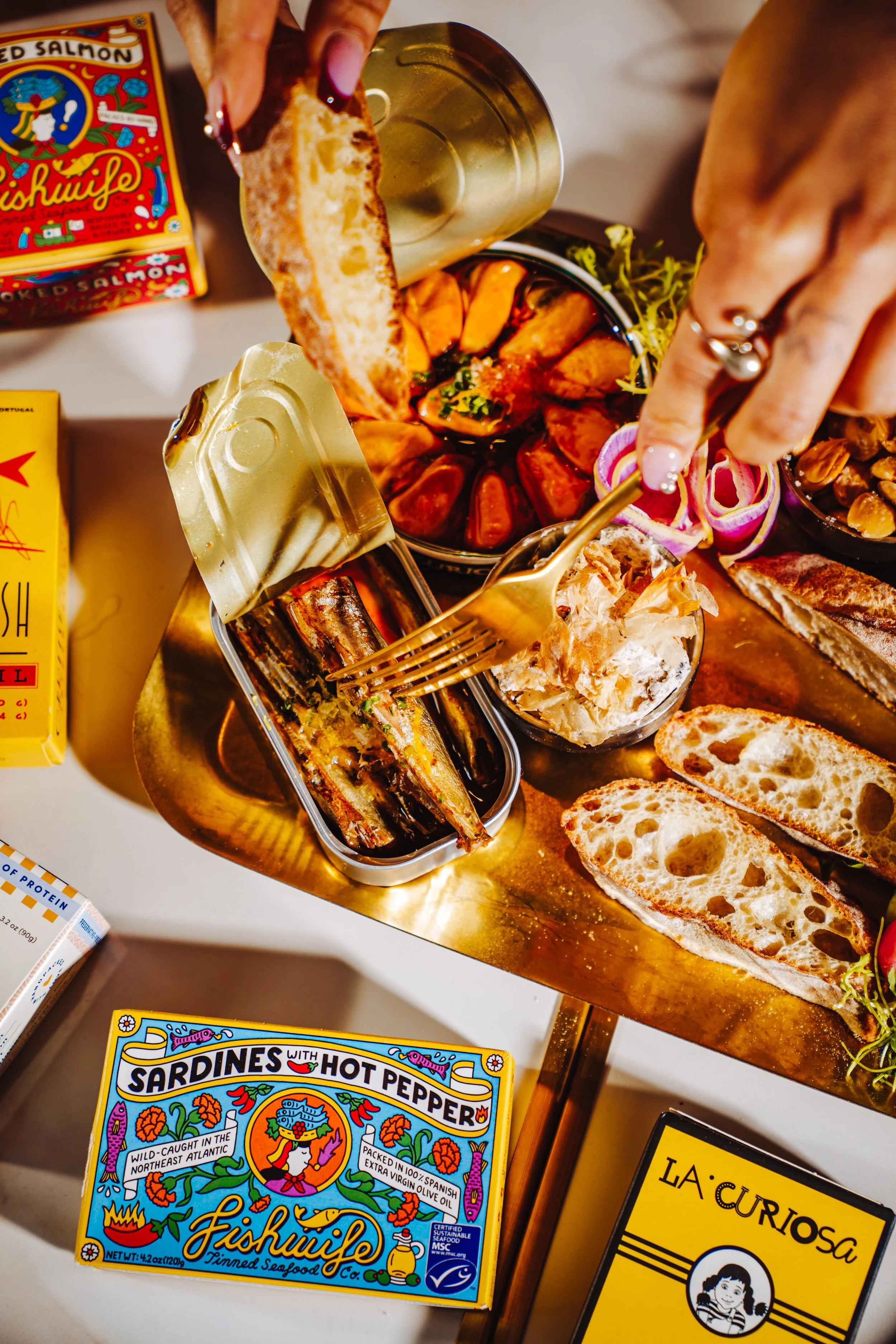 Assorted canned seafood and bread slices on a gold tray, including a box of Fishwife sardines with hot pepper, a pack of La Curiosa canned seafood, and various bowls with roasted vegetables, sliced onions, and other savory items.