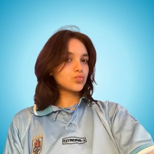 Young woman with shoulder-length brown hair taking a selfie in front of a plain light blue background, wearing a gray sports jersey with text and logos.