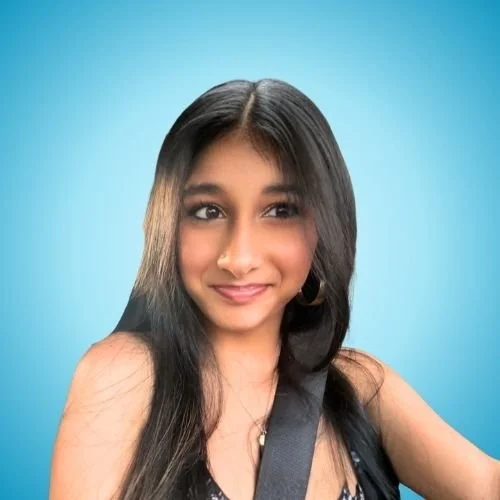 A young woman with long dark hair, wearing a sleeveless top and a seatbelt, smiling against a blue background.